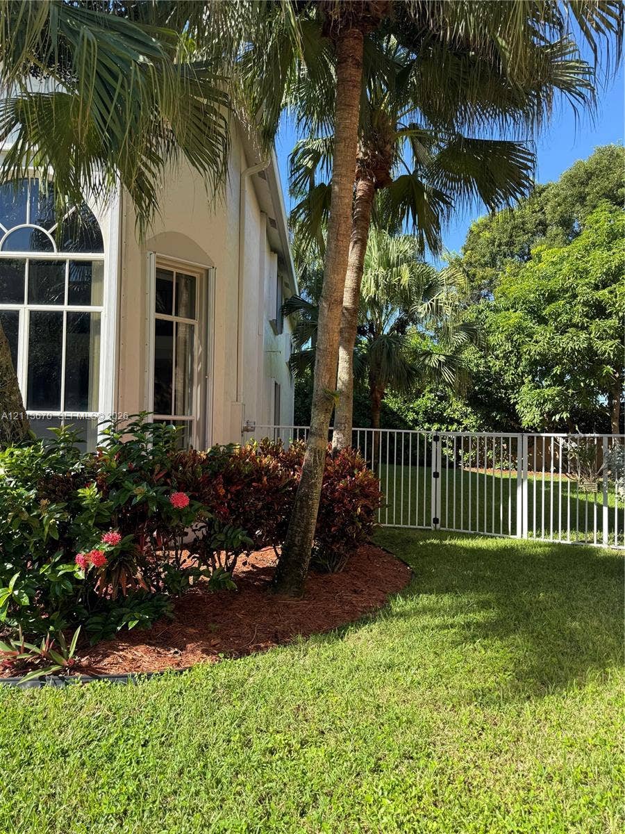 Stucco exterior featuring arched windows, mature palm trees, manicured lawn, and metal perimeter fencing