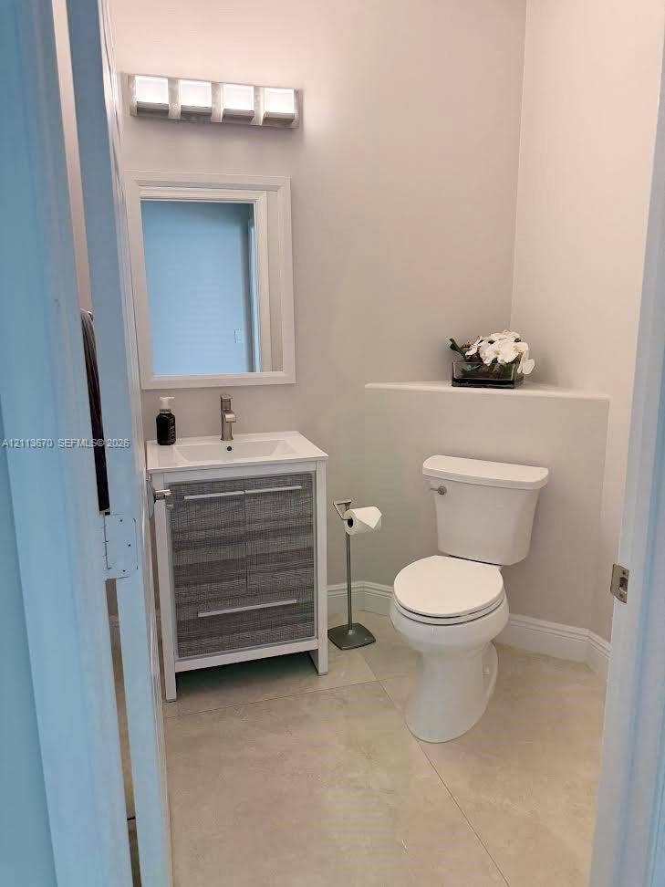 Vanity featuring a textured grey cabinet base, rectangular basin, and single-handle faucet, paired with a white-framed mirror, four-light vanity fixture, and neutral stone-finish tile flooring