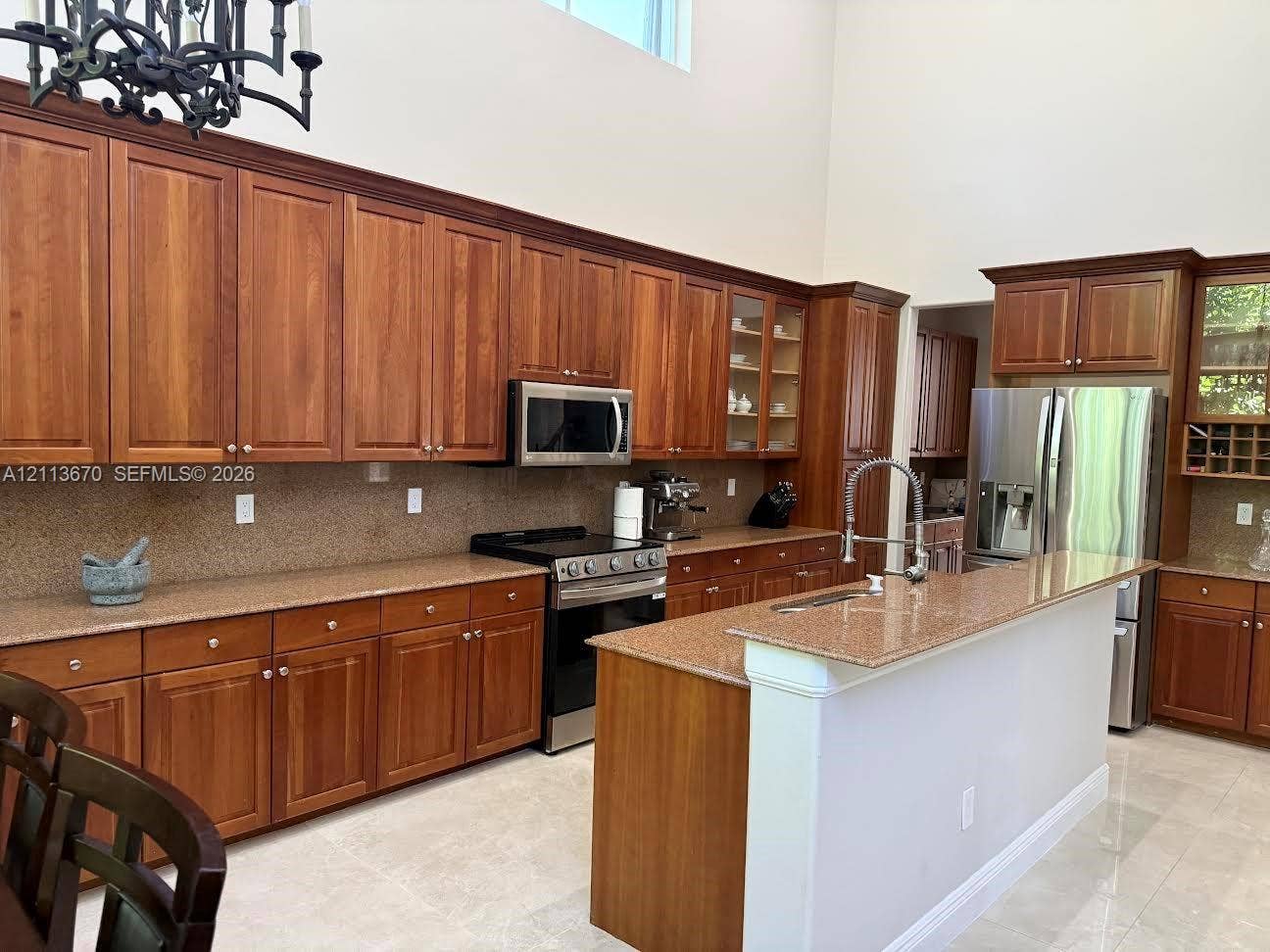 Kitchen featuring stained wood cabinetry, stone-surface countertops, stainless steel appliances, a central island with a sink, and high ceilings