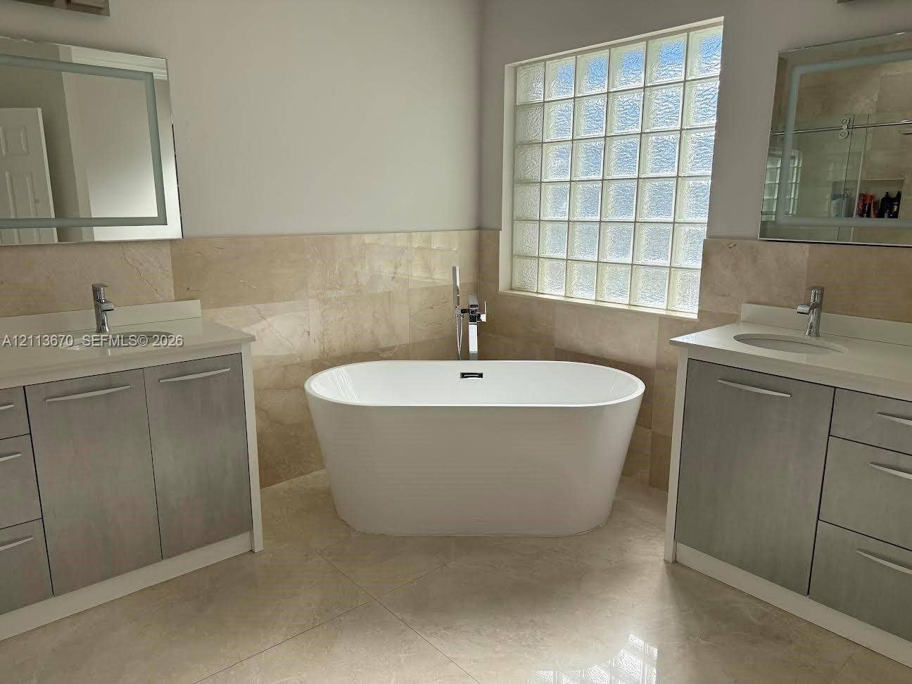 Freestanding oval soaking tub with floor-mounted faucet, dual vanities with integrated sinks, stone-finish tile backsplash, and glass block window