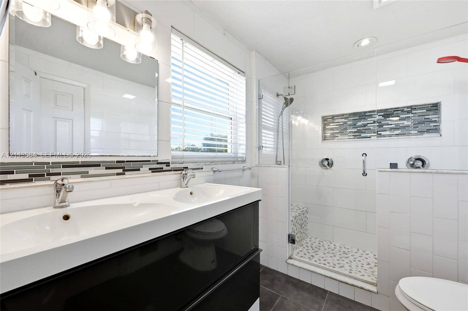 6504 Lenore Drive, Tampa, Tampa, FL 33634 Dual-sink vanity with a white countertop and high-gloss cabinetry, glass-enclosed shower featuring stone tile flooring and decorative inlay, and white subway-style wall tiling