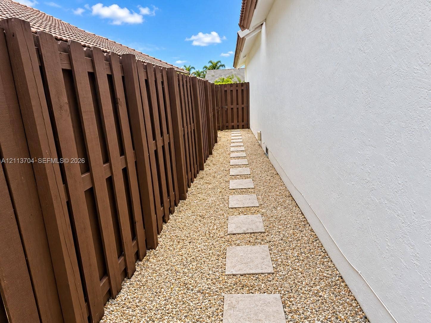 5254 SW 153rd Court, Miami, FL 33185 Exterior side yard featuring a white stucco wall, wood-finish privacy fencing, a decorative stone pathway, and a gravel ground surface