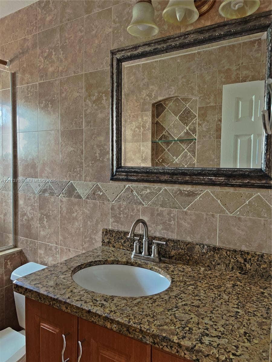 5170 SW 40th Avenue, #1E, Dania, Dania Beach, FL 33314 Vanity featuring a stone-finish countertop with an undermount sink, wood-finish cabinetry, full-height wall tiling with decorative tile inlay, and a built-in recessed wall niche