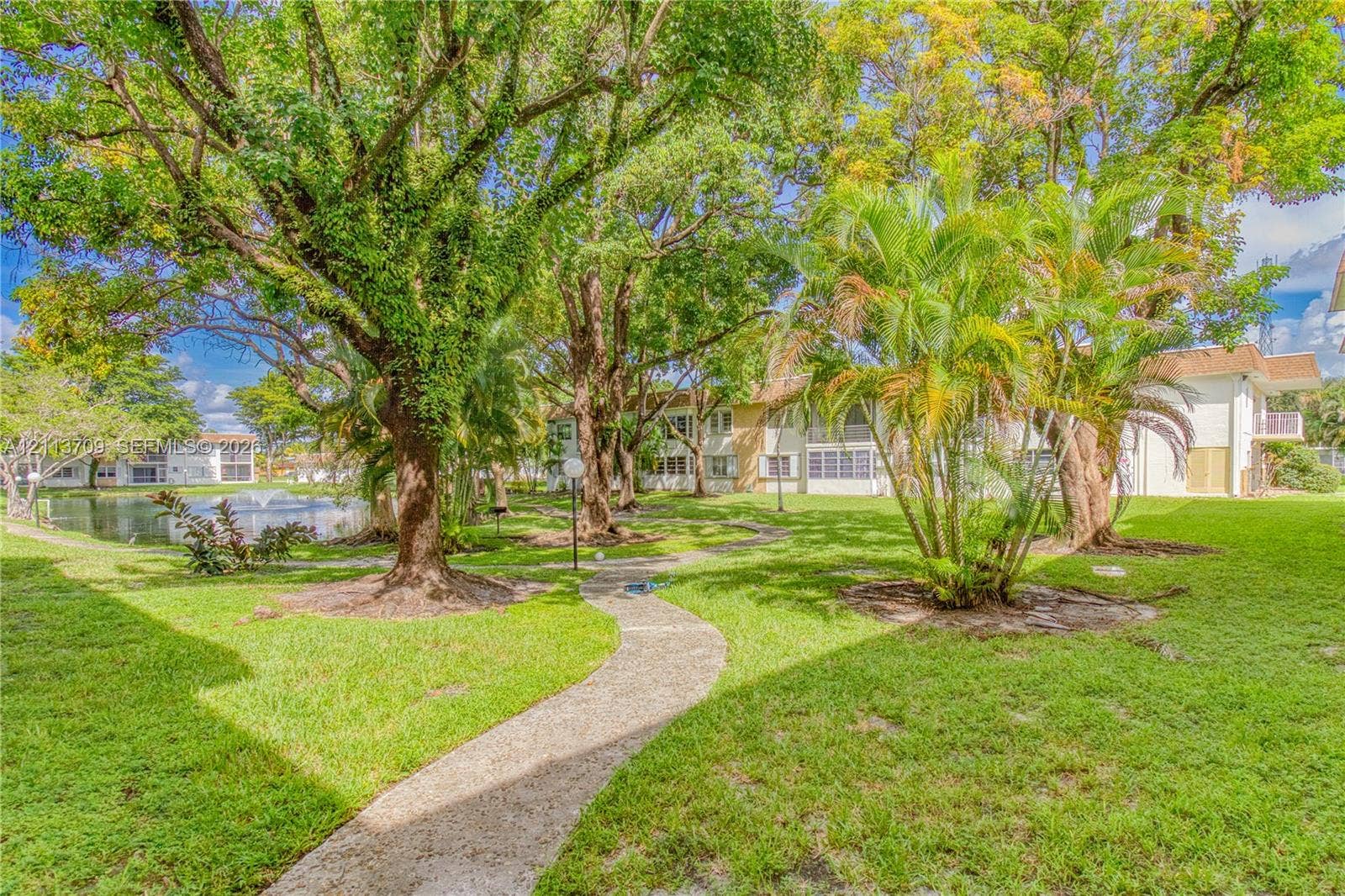 5170 SW 40th Avenue, #1E, Dania, Dania Beach, FL 33314 Landscaped grounds featuring a paved walking path, mature trees, ornamental palms, and a water feature