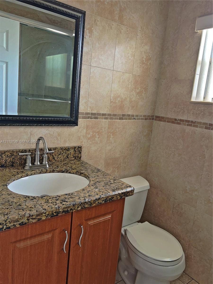 5170 SW 40th Avenue, #1E, Dania, Dania Beach, FL 33314 Bathroom vanity featuring a stone countertop with an undermount sink, wood-finish cabinetry, decorative wall tile with a mosaic accent border, and a wall-mounted framed mirror