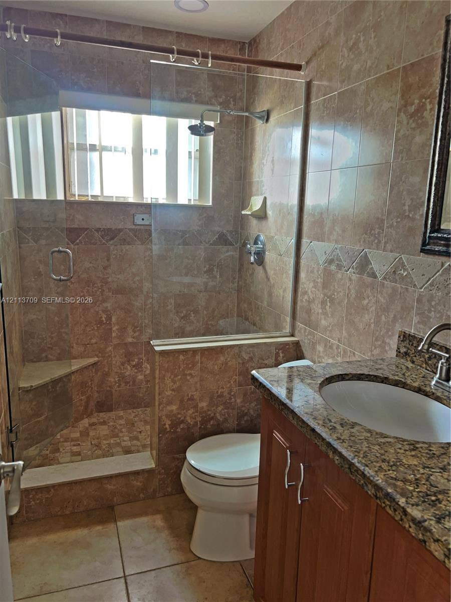 5170 SW 40th Avenue, #1E, Dania, Dania Beach, FL 33314 Bathroom featuring floor-to-ceiling stone-tiled walls, glass-enclosed walk-in shower with integrated corner bench, vanity with stone countertop, and undermount sink