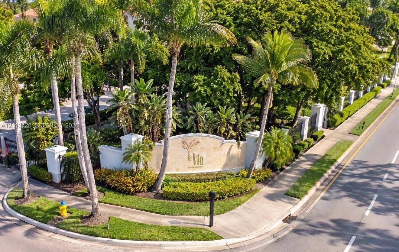 Community entrance monument featuring a stone-finish masonry sign, mature palm trees, manicured hedges, and ornate metal fencing