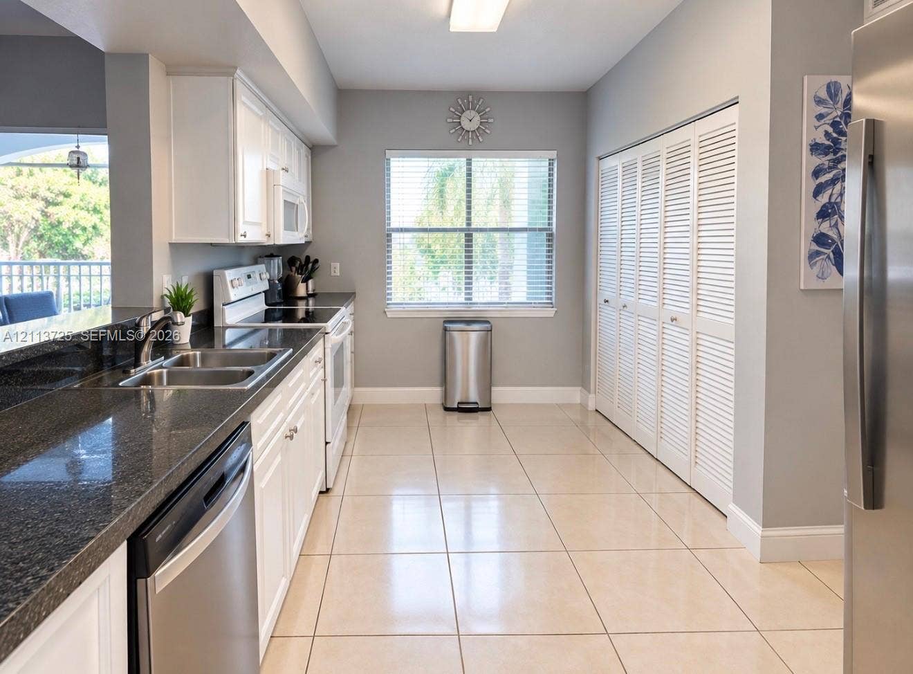Kitchen featuring white shaker-style cabinetry, dark stone-finish countertops, stainless steel appliances, large-format tile flooring, and floor-to-ceiling louvered closet doors
