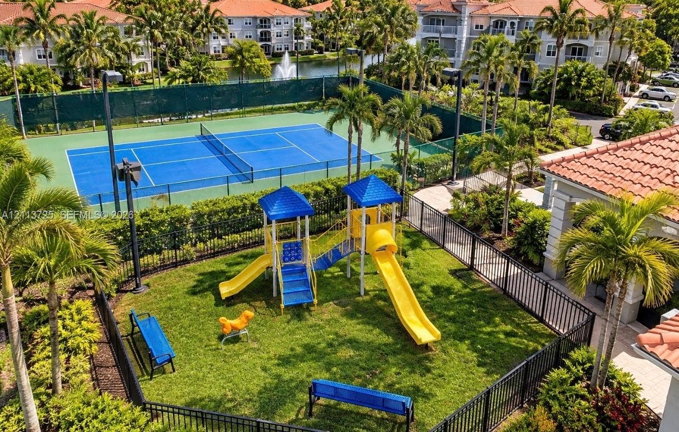 Fenced playground area featuring a jungle gym with slides, a paved tennis court, and lush landscaping with mature palm trees