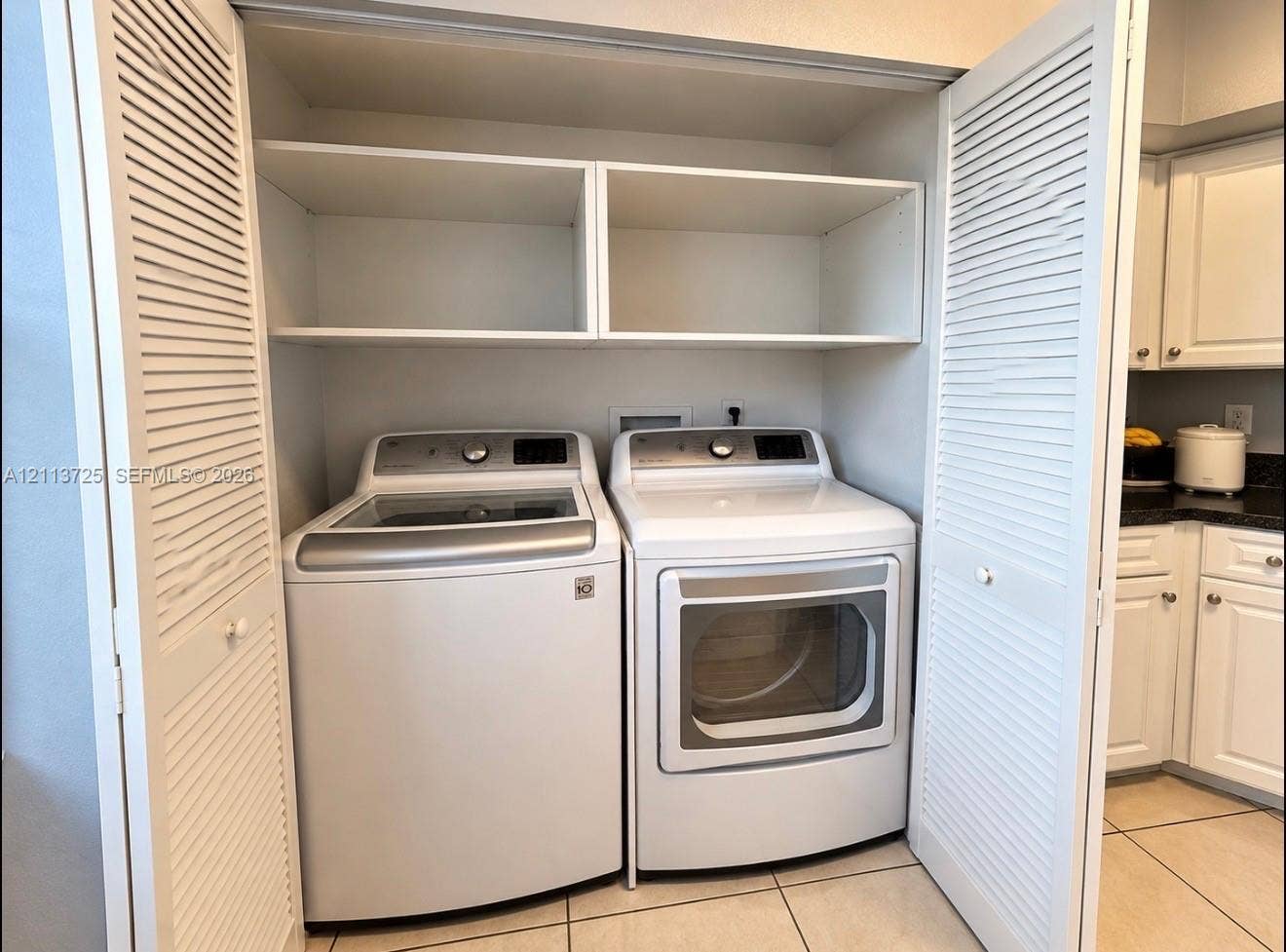 Laundry closet featuring louvered bi-fold doors, white upper shelving, and tiled flooring