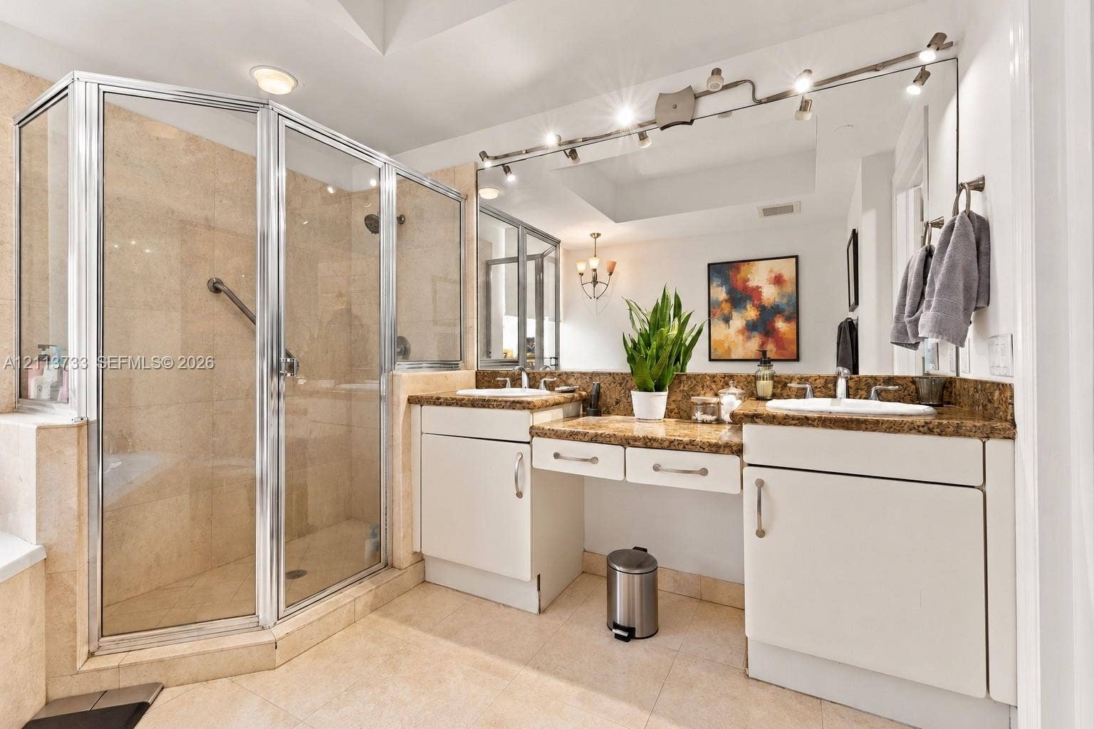 9751 E Bay Harbor Drive, #6A, Bay Harbor Islands, FL 33154 Double vanity featuring stone countertops with dual undermount sinks, central dressing table, full-width mirror with integrated track lighting, and a walk-in glass-enclosed shower with tile surround