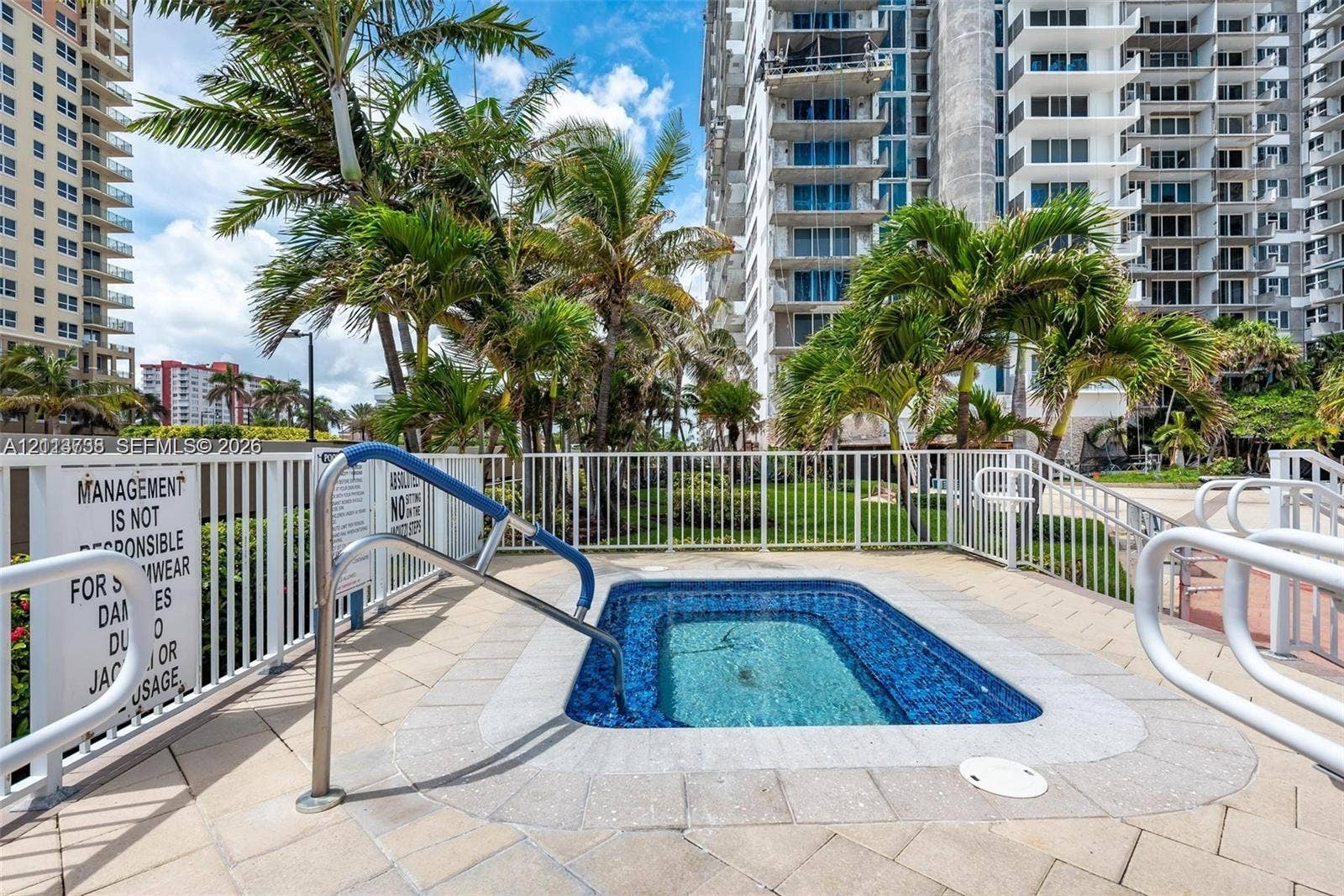 2030 S Ocean Drive, #1719, Hallandale Beach, FL 33009 In-ground spa with tile lining, surrounded by a paved deck, white metal perimeter fencing, and mature palm trees