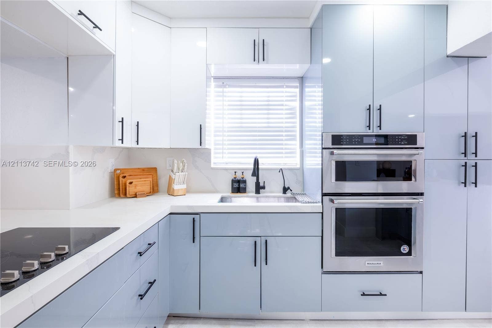 1930 NW 163rd St Road, Miami Gardens, FL 33054 High-gloss cabinetry featuring white and pale blue finishes, solid surface countertops with integrated backsplash, built-in stainless steel double wall ovens, black hardware, and an undermount sink with a high-arch matte black faucet
