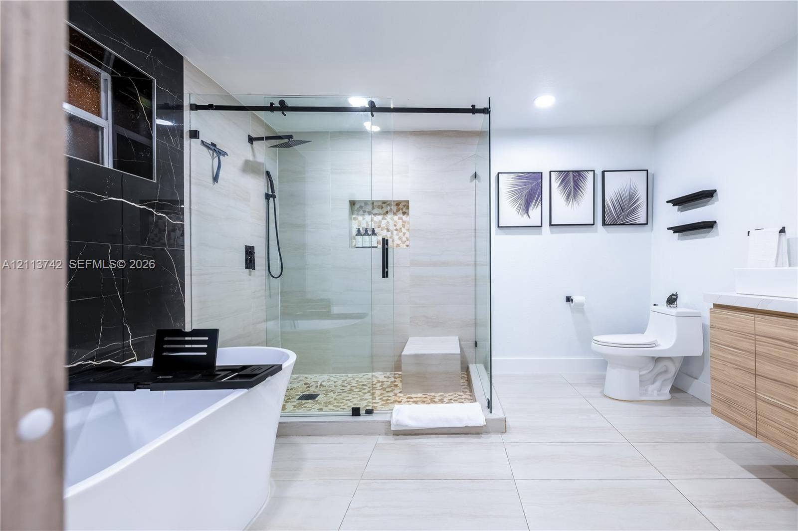 1930 NW 163rd St Road, Miami Gardens, FL 33054 Freestanding soaking tub, glass-enclosed walk-in shower with stone tile flooring, built-in shower niche, wall-mounted wood-finish vanity, and floor-to-ceiling contrasting wall tile