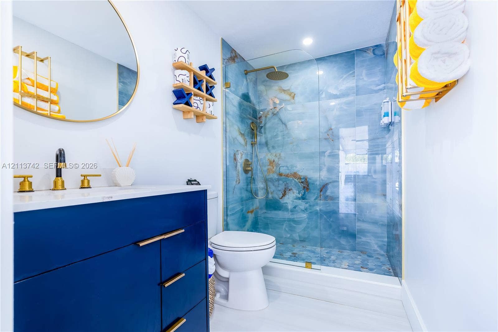 1930 NW 163rd St Road, Miami Gardens, FL 33054 Blue vanity featuring a white countertop and gold-tone hardware, oval gold-framed mirror, and a walk-in shower with floor-to-ceiling patterned wall tile, a frameless glass door, and a gold-tone rainfall showerhead