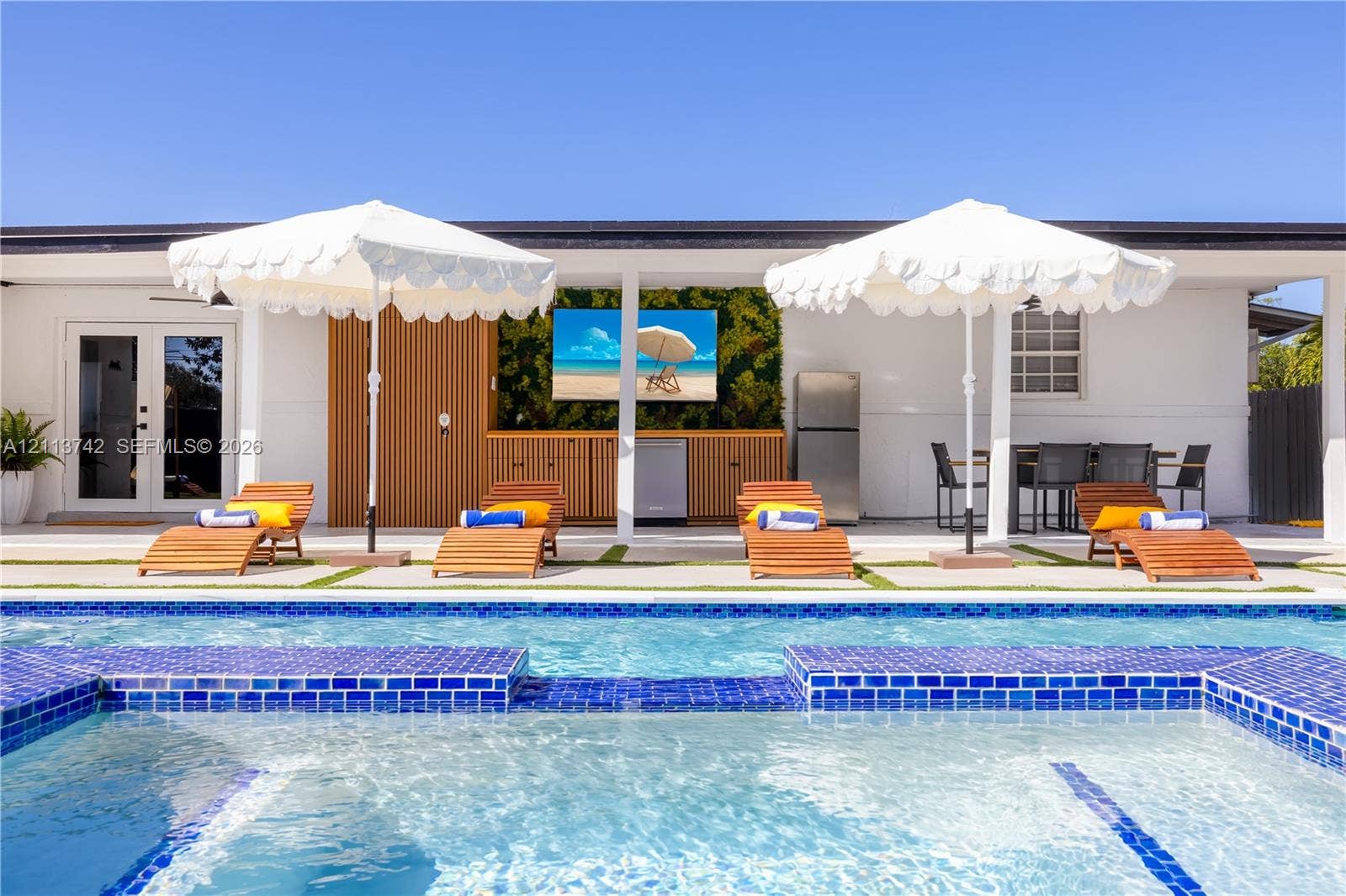 1930 NW 163rd St Road, Miami Gardens, FL 33054 Outdoor swimming pool with blue tile edging, paired with a covered patio featuring built-in wood-slat cabinetry and a recessed television mounting area
