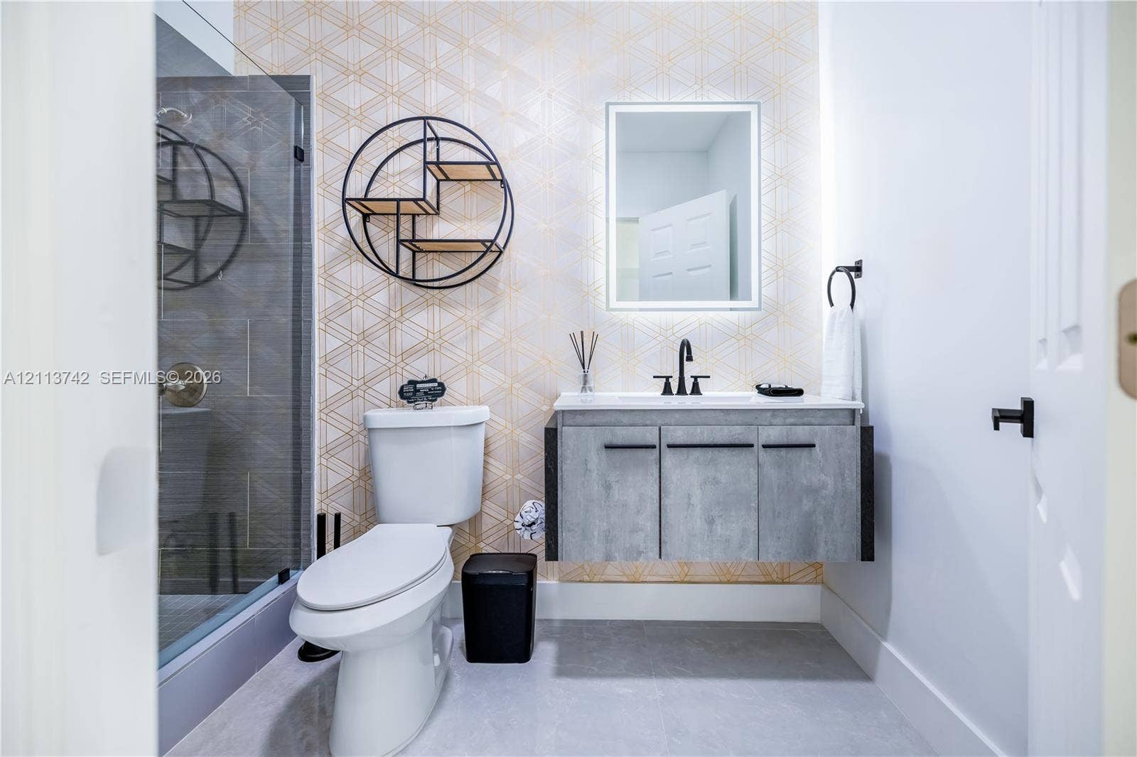 1930 NW 163rd St Road, Miami Gardens, FL 33054 Floating vanity with a concrete-finish cabinet and solid surface countertop, geometric patterned wall covering, glass-enclosed shower with tiled surround, and LED-backlit mirror