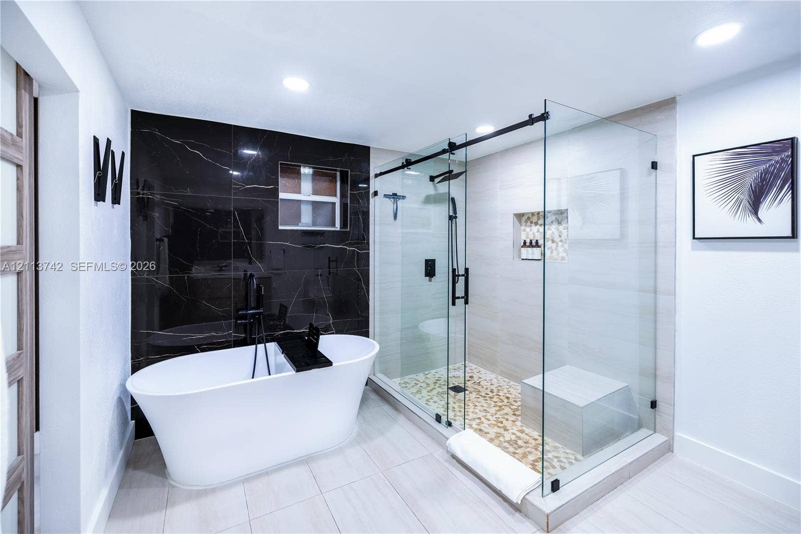 1930 NW 163rd St Road, Miami Gardens, FL 33054 Freestanding soaking tub with matte black floor-mounted faucet, walk-in glass-enclosed shower with built-in bench, pebble-style shower flooring, recessed wall niche, and contrasting black tile accent wall