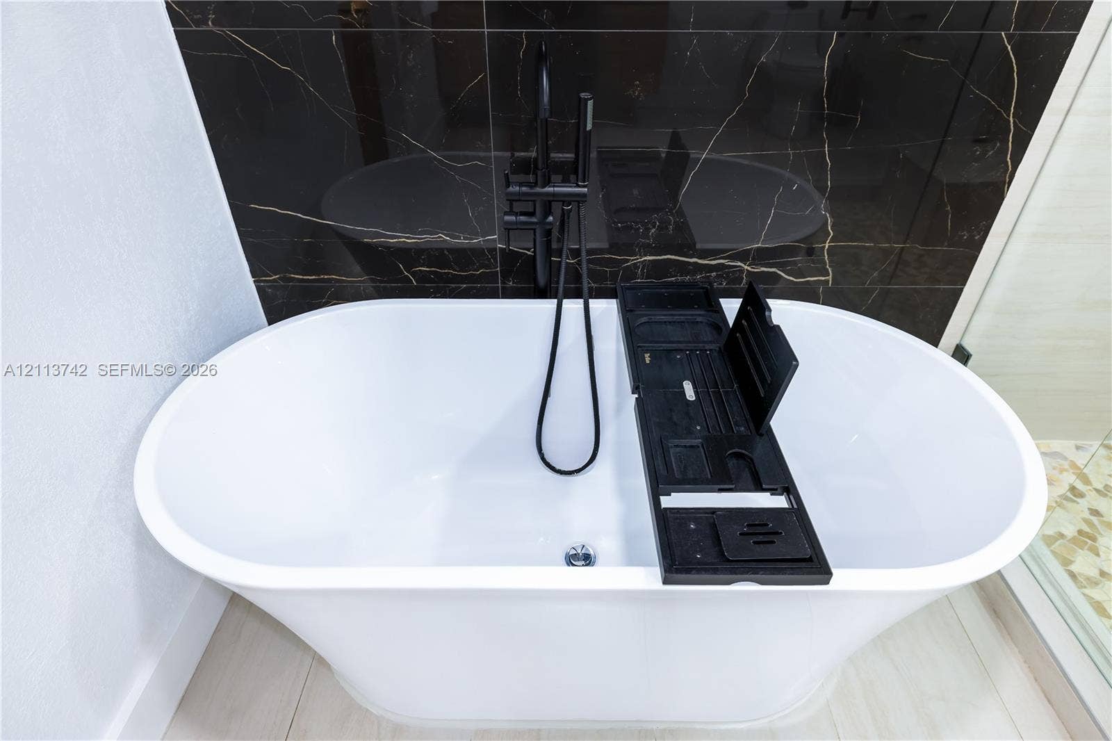 1930 NW 163rd St Road, Miami Gardens, FL 33054 Freestanding white soaking tub with a floor-mounted matte black faucet and handheld shower attachment, set against a dark, veined tile accent wall