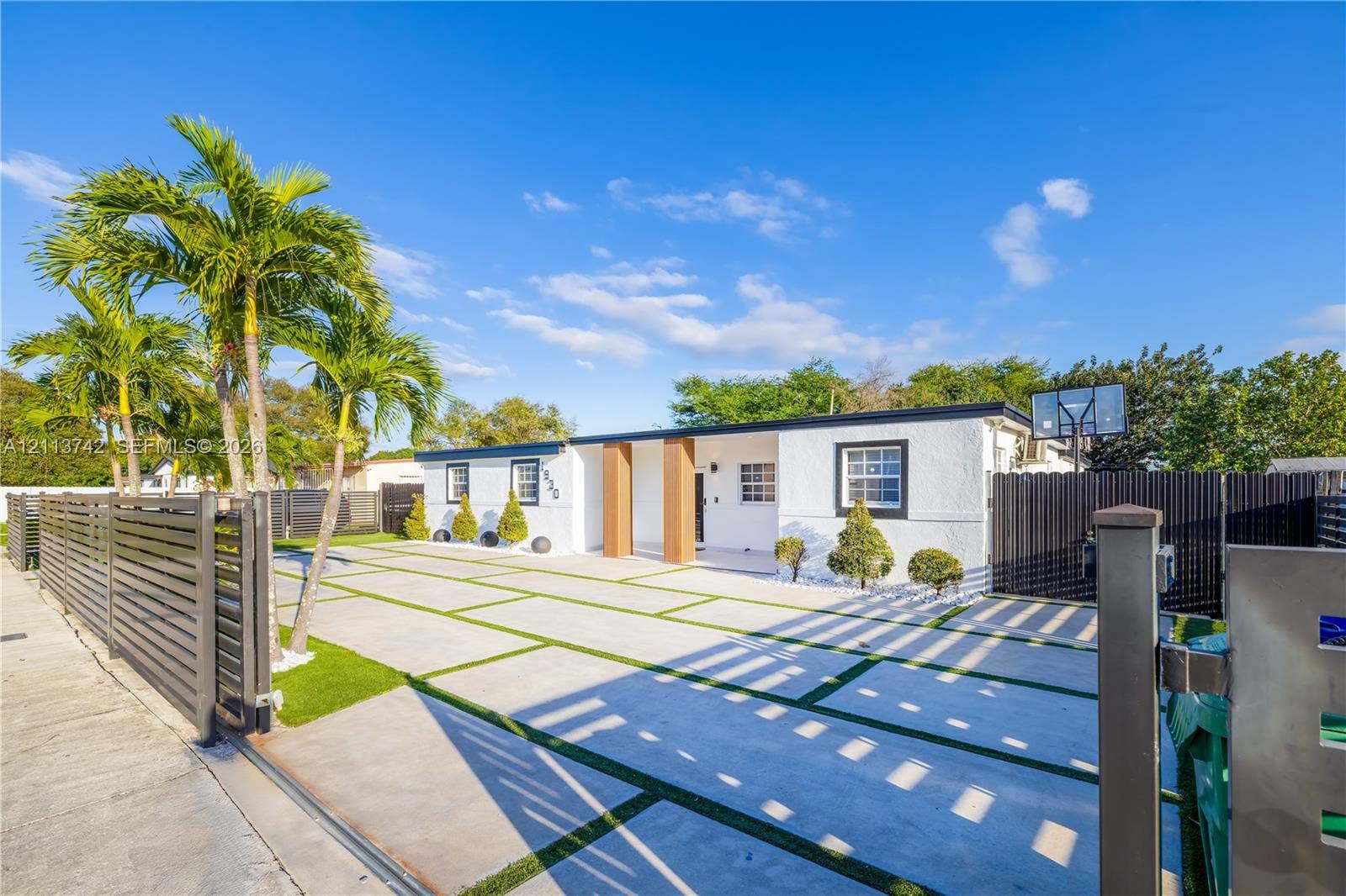 1930 NW 163rd St Road, Miami Gardens, FL 33054 Single-story exterior featuring white stucco siding, a concrete paver driveway with turf accents, mature palm trees, and perimeter horizontal slat fencing