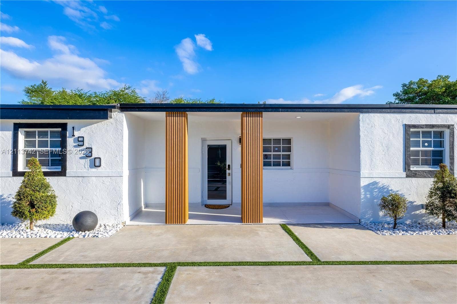 1930 NW 163rd St Road, Miami Gardens, FL 33054 Modern exterior featuring a white stucco facade, a covered entryway with vertical wood-slat columns, and a grid-patterned concrete driveway with integrated turf strips