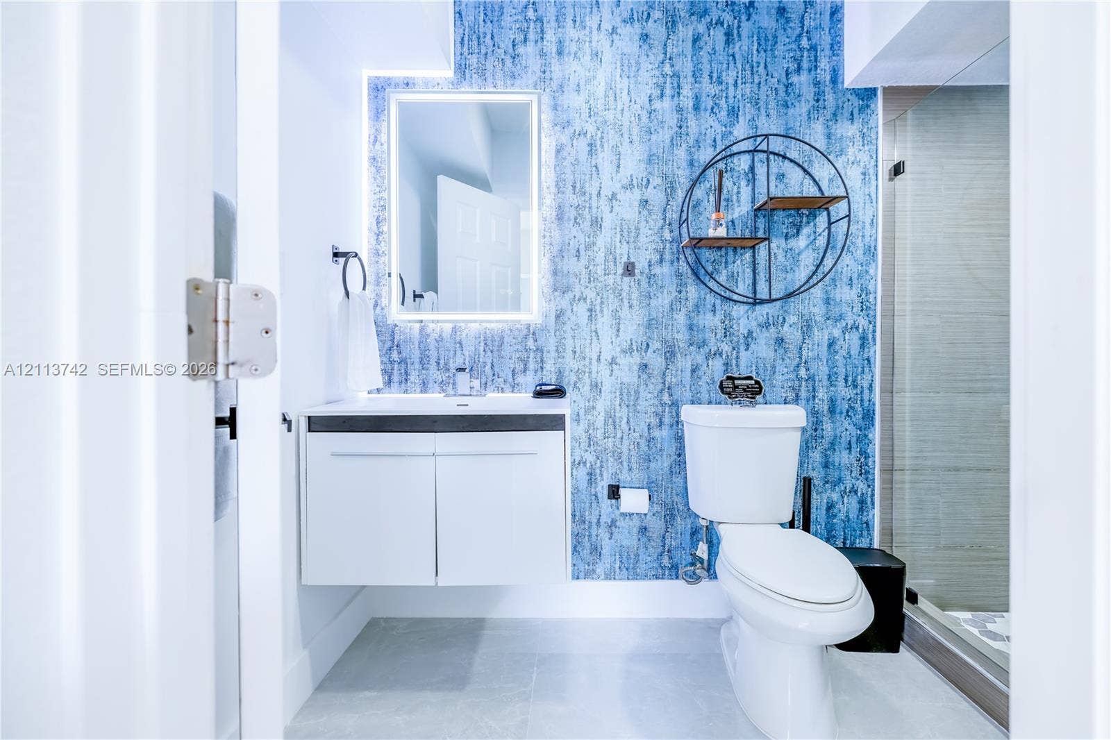 1930 NW 163rd St Road, Miami Gardens, FL 33054 Bathroom featuring a wall-mounted floating vanity, backlit rectangular mirror, textured blue accent wall, and glass-enclosed shower