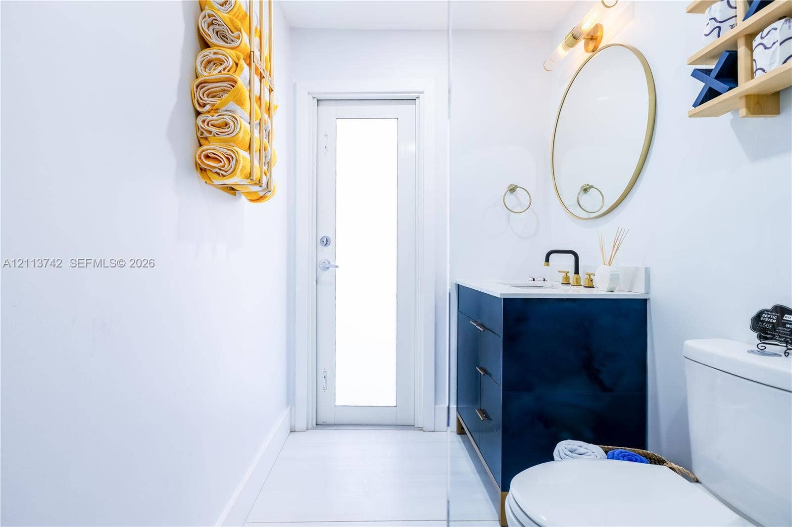 1930 NW 163rd St Road, Miami Gardens, FL 33054 Bathroom featuring a dark blue vanity with gold hardware, an oval mirror with a gold frame, a modern wall-mounted light fixture, and a door with an inset glass panel