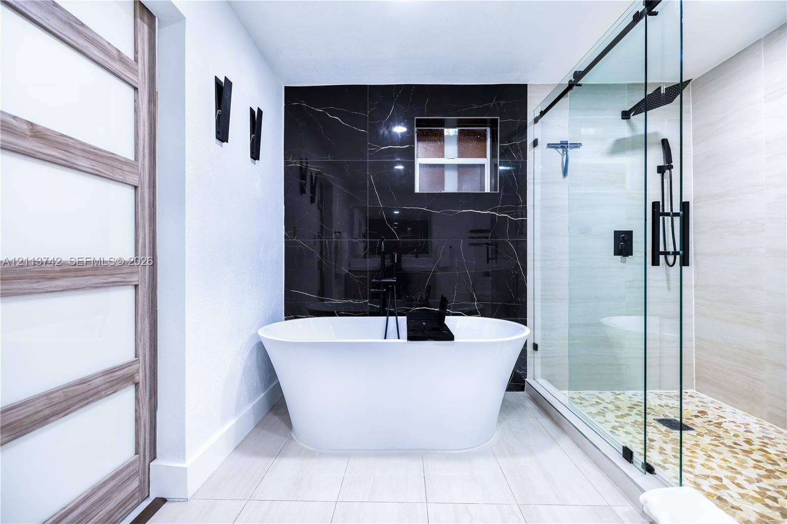1930 NW 163rd St Road, Miami Gardens, FL 33054 Freestanding soaking tub, walk-in glass-enclosed shower with pebble-style floor tiling, dark stone-finish accent wall, and wood-finish door with frosted glass panels