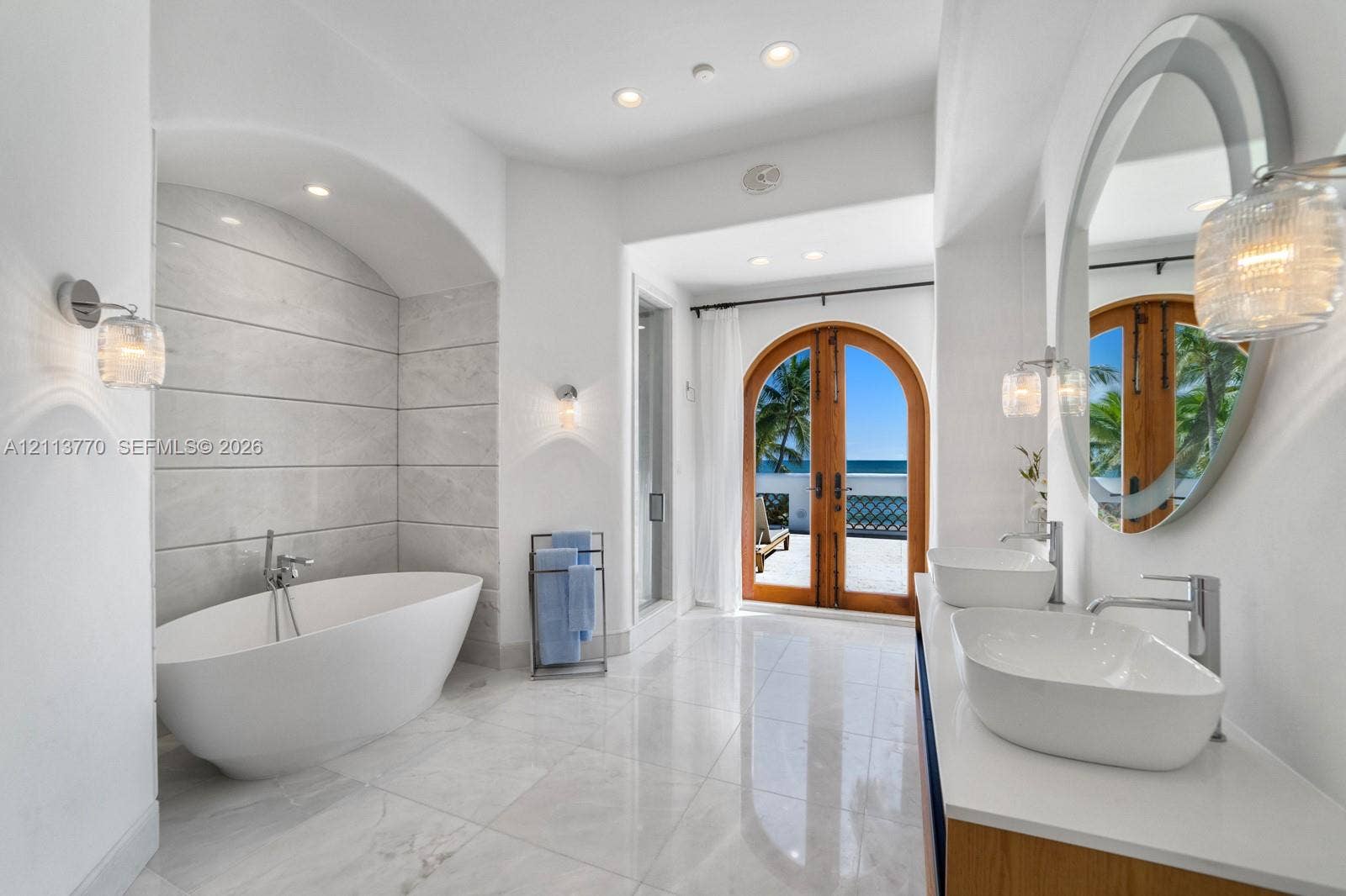 387 Ocean Boulevard, Golden Beach, Golden Beach, FL 33160 Freestanding soaking tub set within a tiled arched alcove, dual oval vessel sinks with polished chrome faucets, quartz countertop vanity, and arched double doors featuring exterior ocean scenery