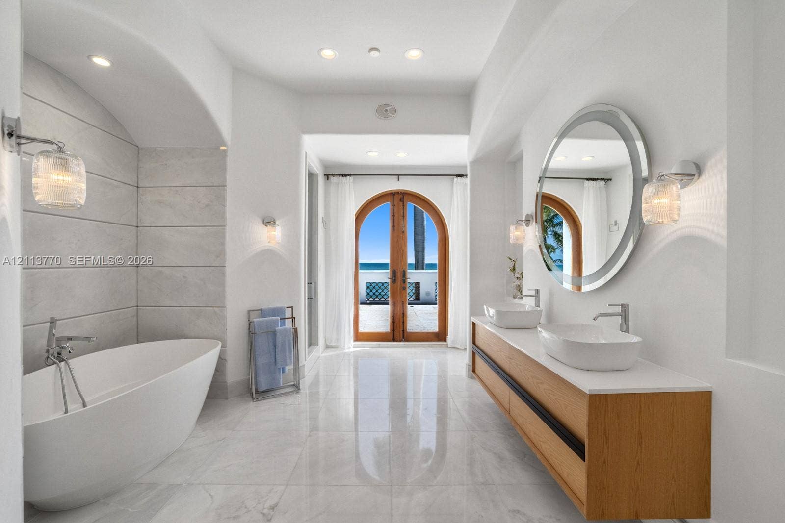 387 Ocean Boulevard, Golden Beach, Golden Beach, FL 33160 Freestanding soaking tub set within a tiled arched alcove, dual-sink floating vanity with white surface, round wall-mounted mirror, recessed lighting, and arched wood-frame glass doors