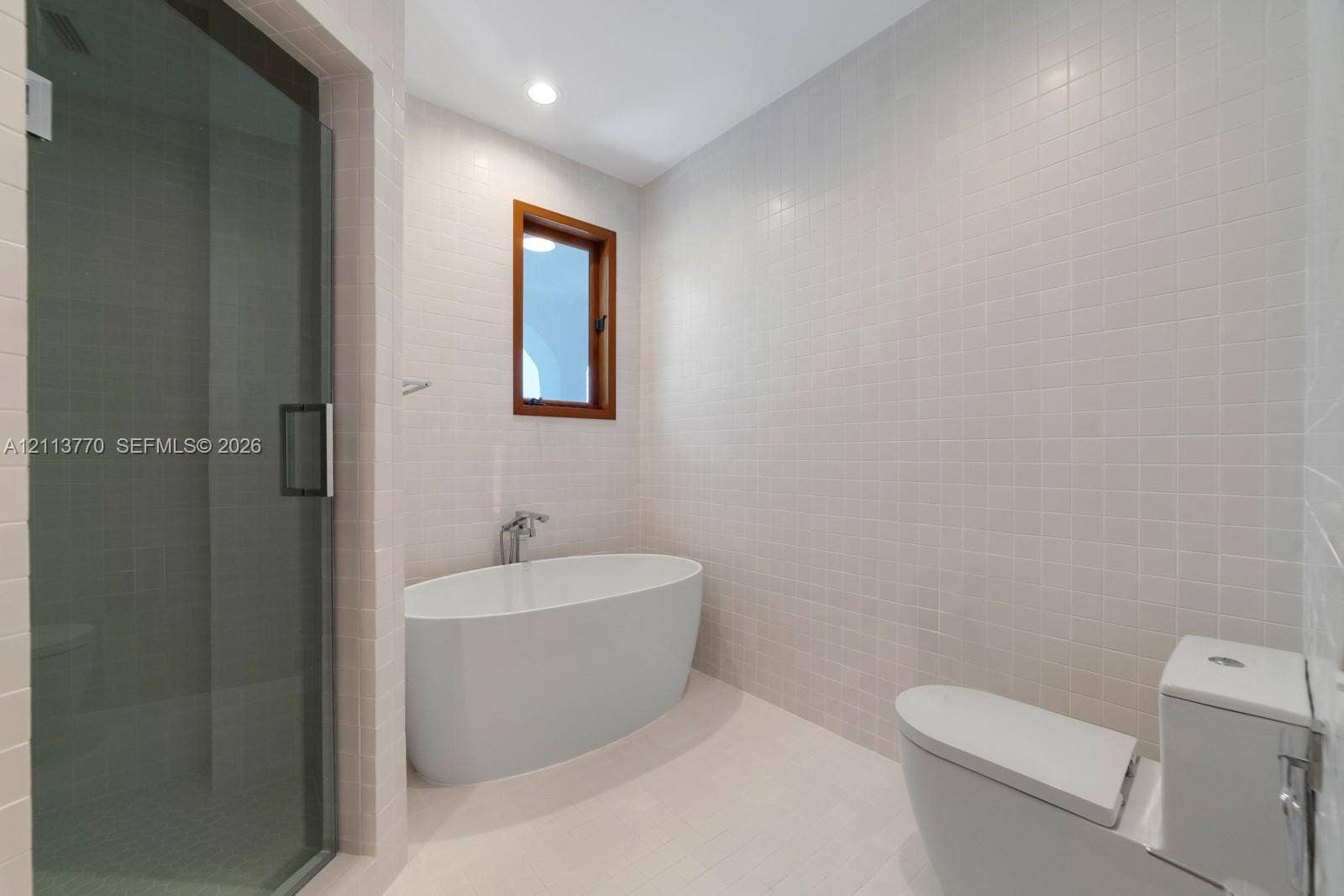 387 Ocean Boulevard, Golden Beach, Golden Beach, FL 33160 Freestanding soaking tub with wall-mounted faucet, glass-enclosed walk-in shower, floor-to-ceiling white tile surround, and wood-framed window