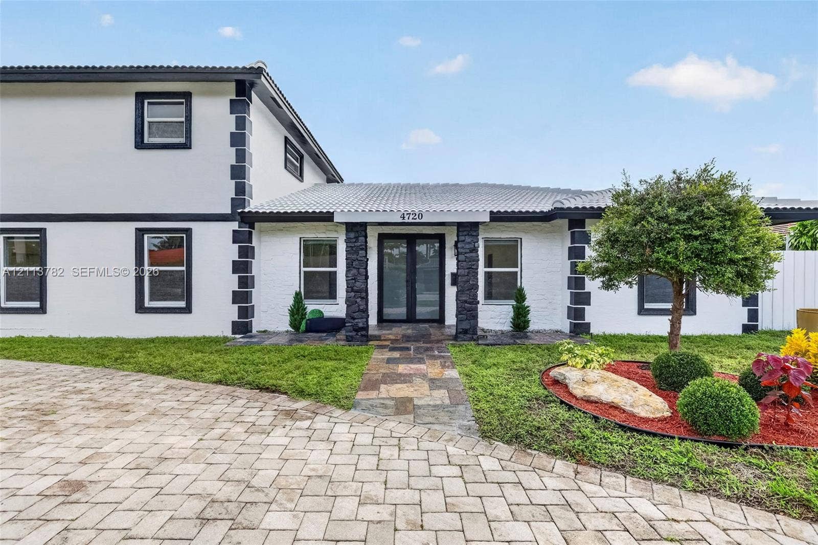 4720 Bayview Drive, Fort Lauderdale, FL 33308 Two-story exterior featuring a white-painted brick facade with dark contrast trim, stone-clad porch columns, a multi-colored stone paver walkway, and a herringbone-patterned concrete paver driveway