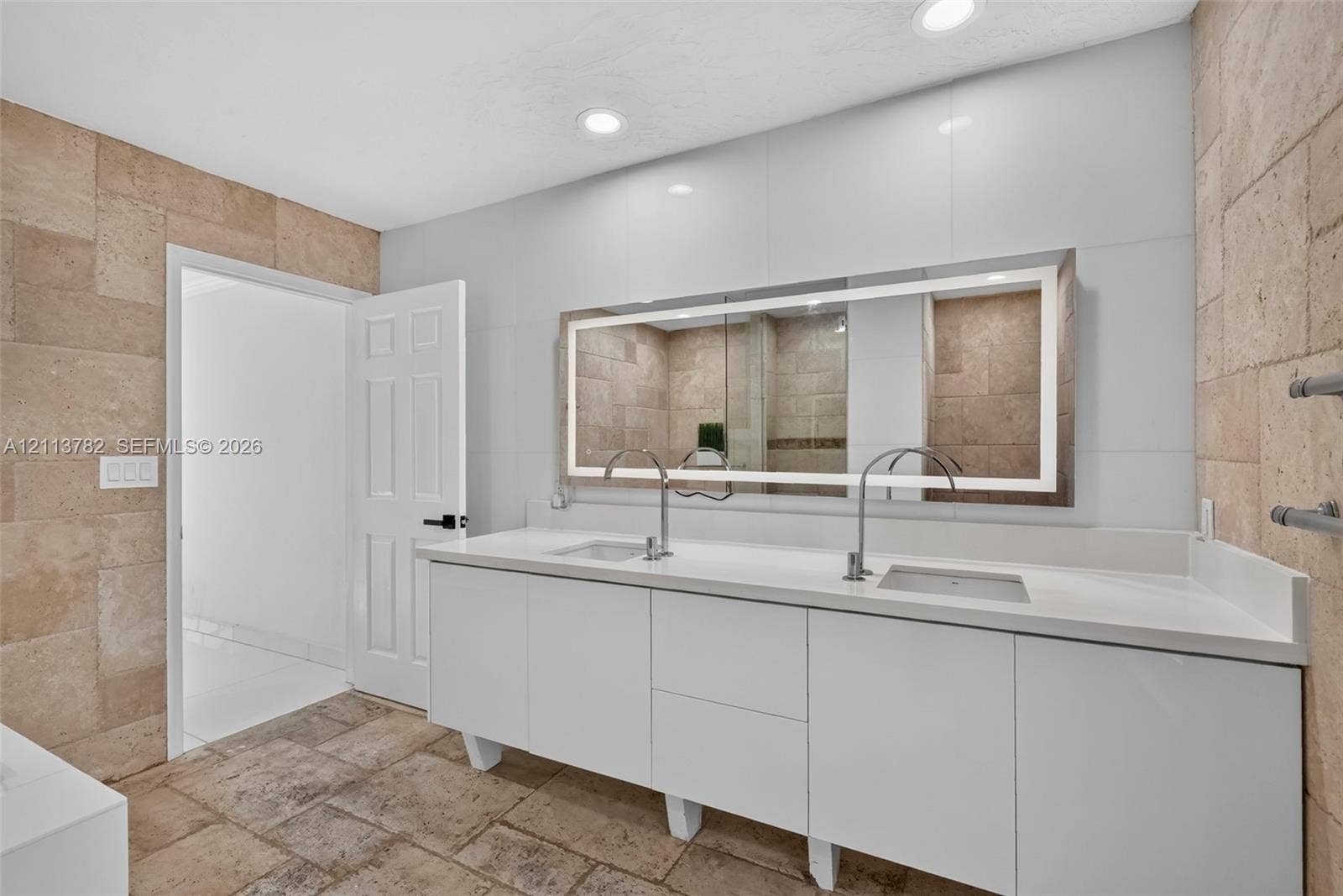 4720 Bayview Drive, Fort Lauderdale, FL 33308 Double vanity with white cabinetry, solid surface countertop, rectangular undermount sinks, tall polished metal faucets, and a large backlit wall-mounted mirror surrounded by textured stone wall tile