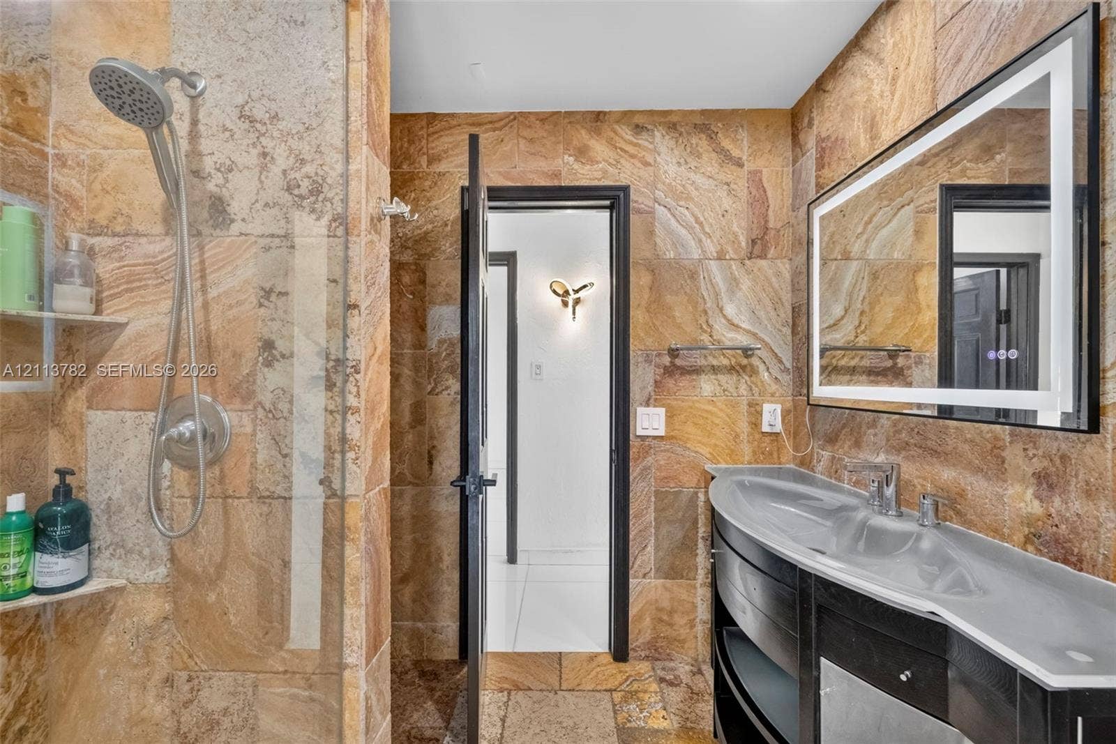 4720 Bayview Drive, Fort Lauderdale, FL 33308 Bathroom featuring natural stone tile wall and floor surfaces, a curved vanity with a basin sink, an integrated-lighting rectangular mirror, and a wall-mounted shower head