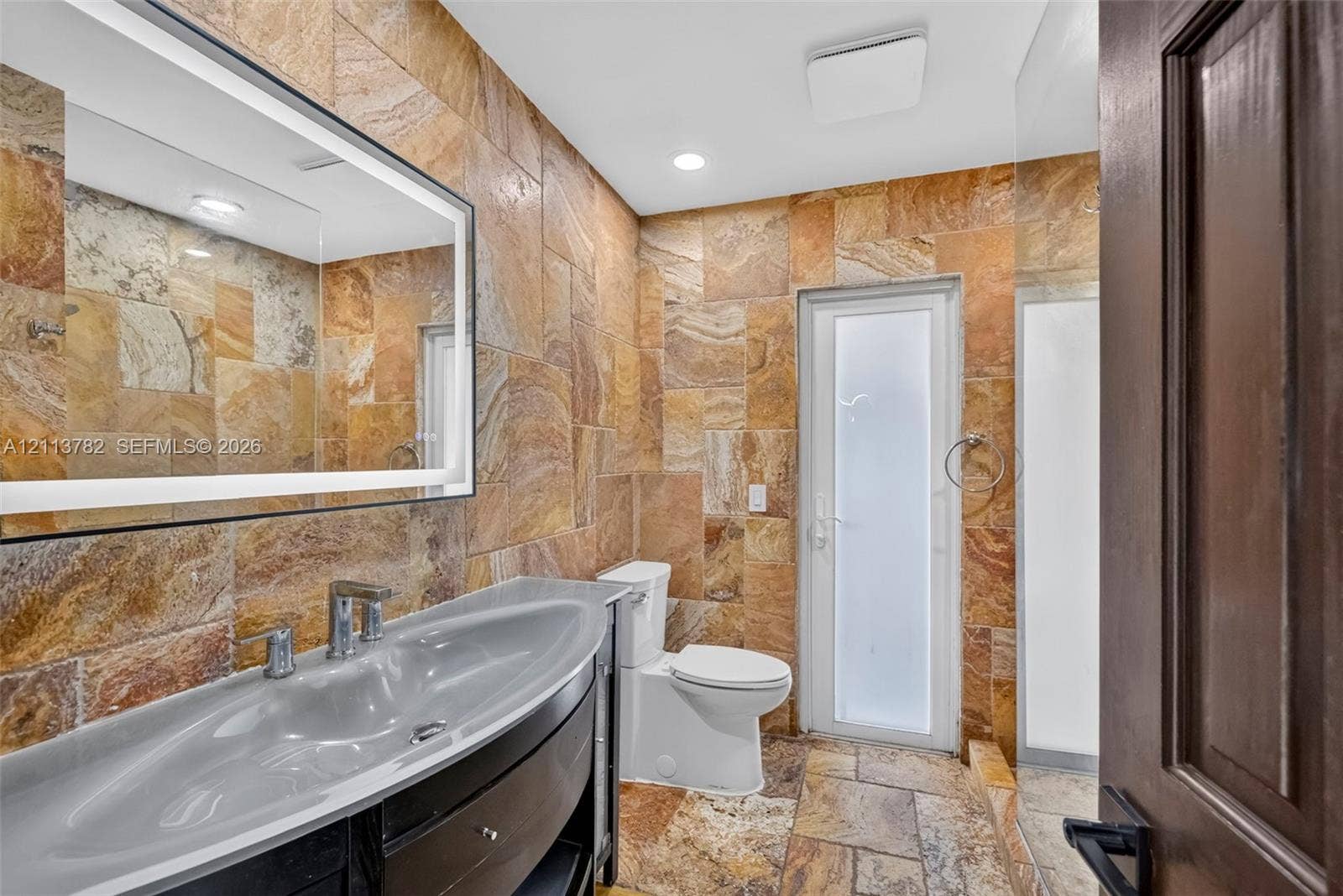 4720 Bayview Drive, Fort Lauderdale, FL 33308 Stone tile wall and floor surfaces, custom vanity with integrated basin, LED-backlit mirror, and frosted glass paneled door