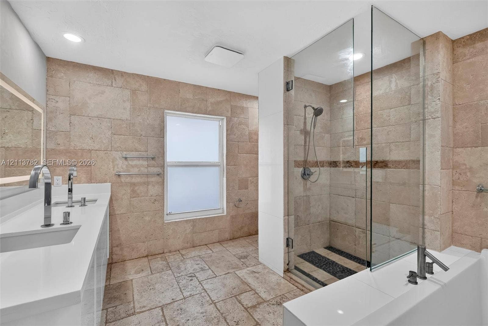 4720 Bayview Drive, Fort Lauderdale, FL 33308 Bathroom featuring natural stone wall and floor tiling, double-basin vanity with solid-surface countertop, glass-enclosed walk-in shower with rainfall showerhead, and a deep soaking tub
