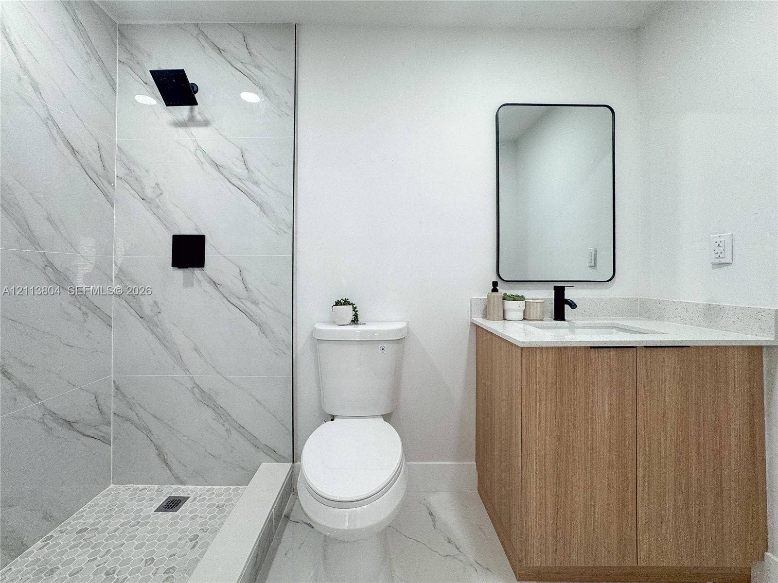 2300 NW 31st Avenue, Fort Lauderdale, FL 33311 Full-height shower surround with veined stone-finish tile, pebble-tile shower floor, matte black shower fixture, light wood-finish vanity with stone-finish countertop, and wall-mounted rectangular mirror with a black frame