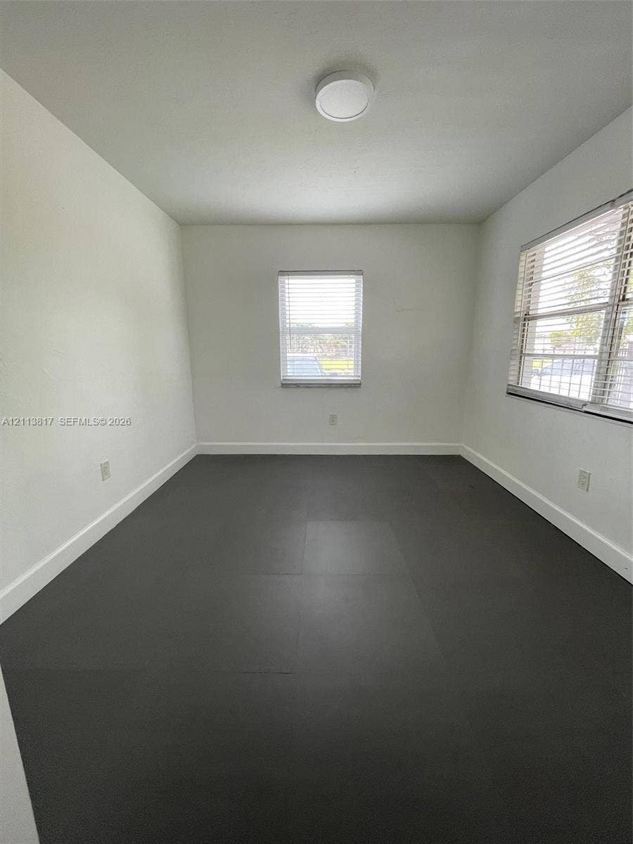 2515 NW 52nd Street, #1, Miami, Miami, FL 33142 Neutral-toned interior space featuring dark grey tile flooring, bright white walls, two multi-pane windows with horizontal blinds, and a flush-mount ceiling light fixture