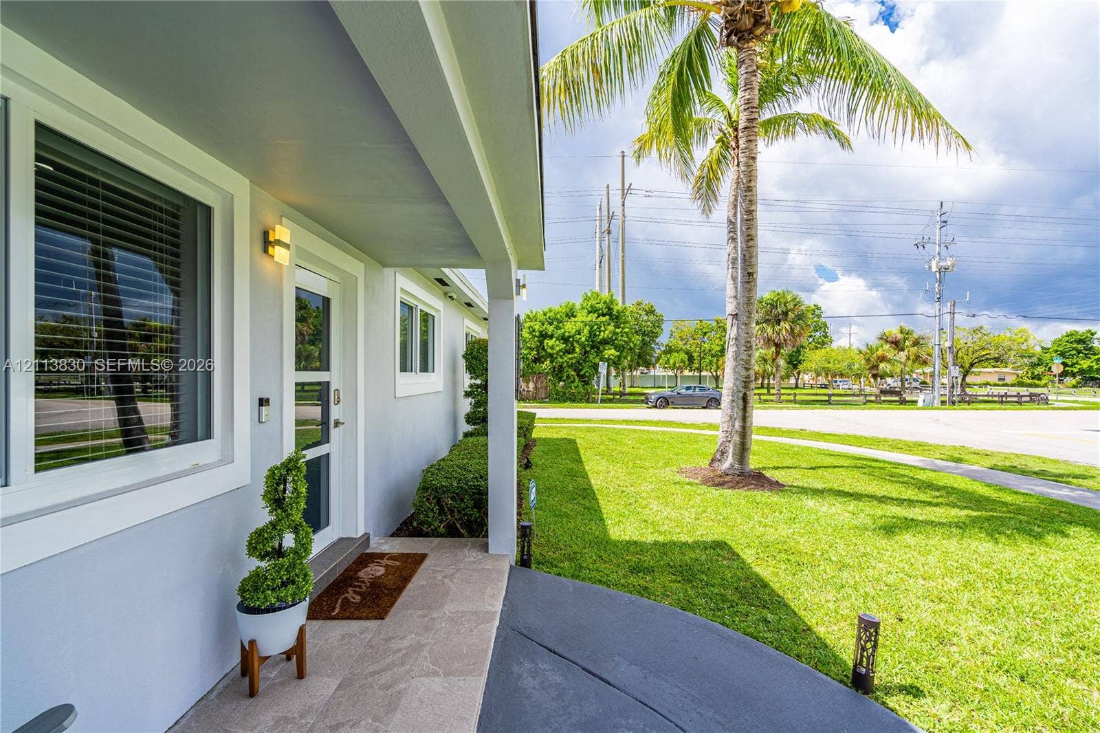 3545 SW 104th Court, Miami, FL 33165 Covered entryway featuring stone-finish floor tiling, exterior wall-mounted lighting, and a glass-paneled entry door, complemented by a manicured lawn and mature palm tree