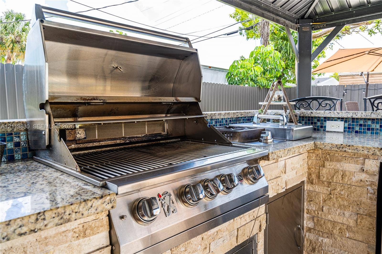 3545 SW 104th Court, Miami, FL 33165 Outdoor kitchen featuring a built-in stainless steel grill, stone veneer base, patterned glass tile backsplash, and stone countertop under a covered structure