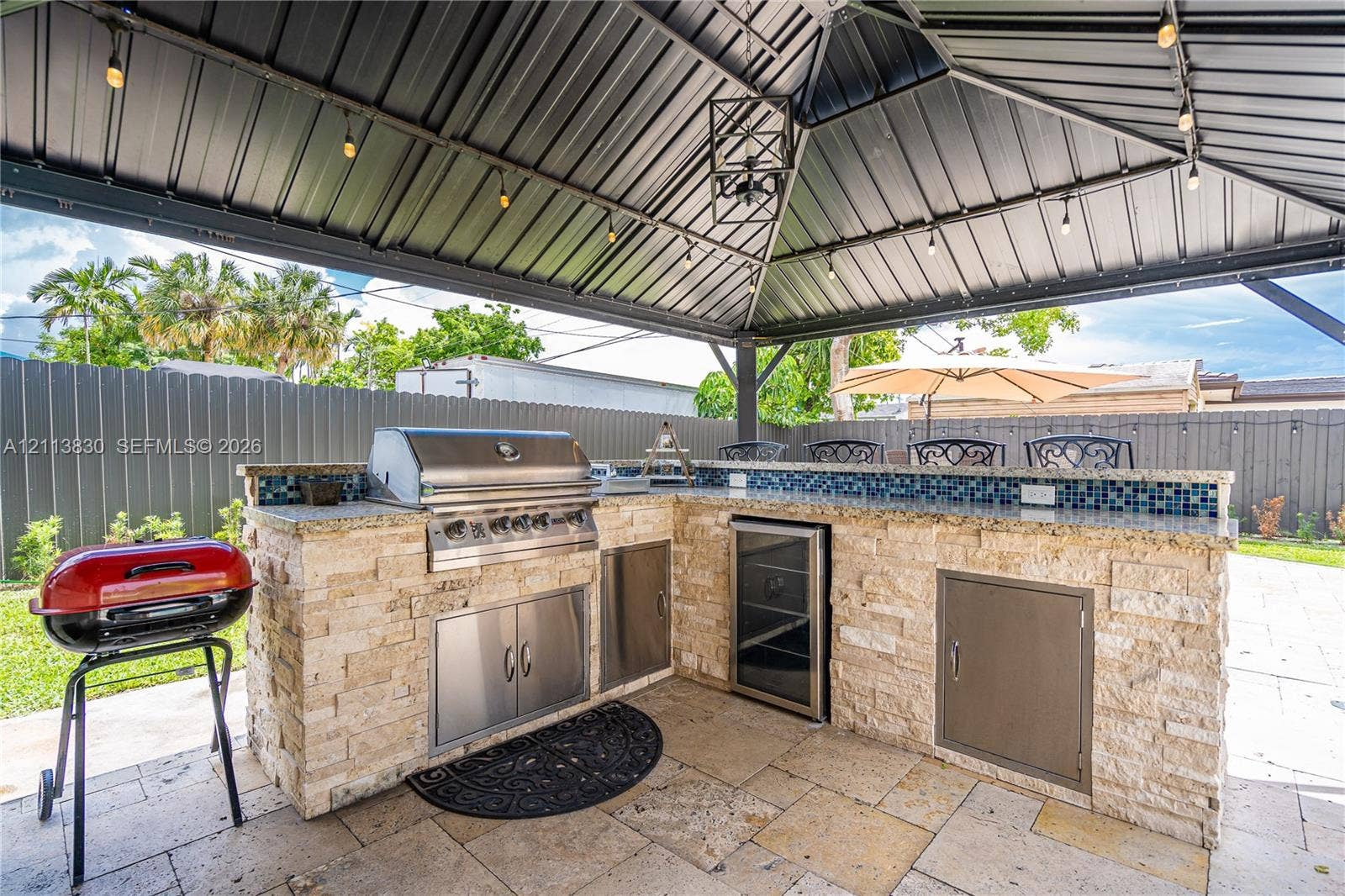 3545 SW 104th Court, Miami, FL 33165 Covered outdoor kitchen featuring a stone-clad island, stainless steel grill, beverage refrigerator, tile backsplash, and dark metal gazebo structure