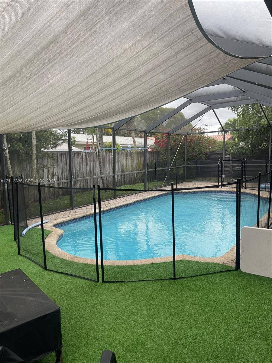 10153 SW 51st Street, Cooper City, Cooper City, FL 33328 Screened-in pool area featuring a brick paver surround, synthetic turf ground cover, removable black safety fencing, and an overhead sun shade