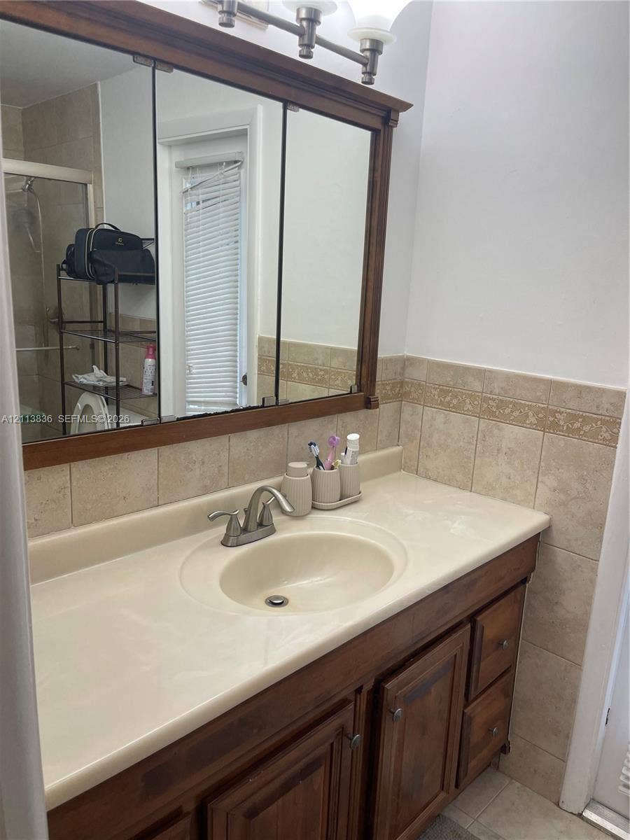 10153 SW 51st Street, Cooper City, Cooper City, FL 33328 Bathroom vanity featuring wood-finish cabinetry, a molded countertop with integrated basin, polished metal faucet, tri-fold medicine cabinet, and tiled backsplash with decorative inlay