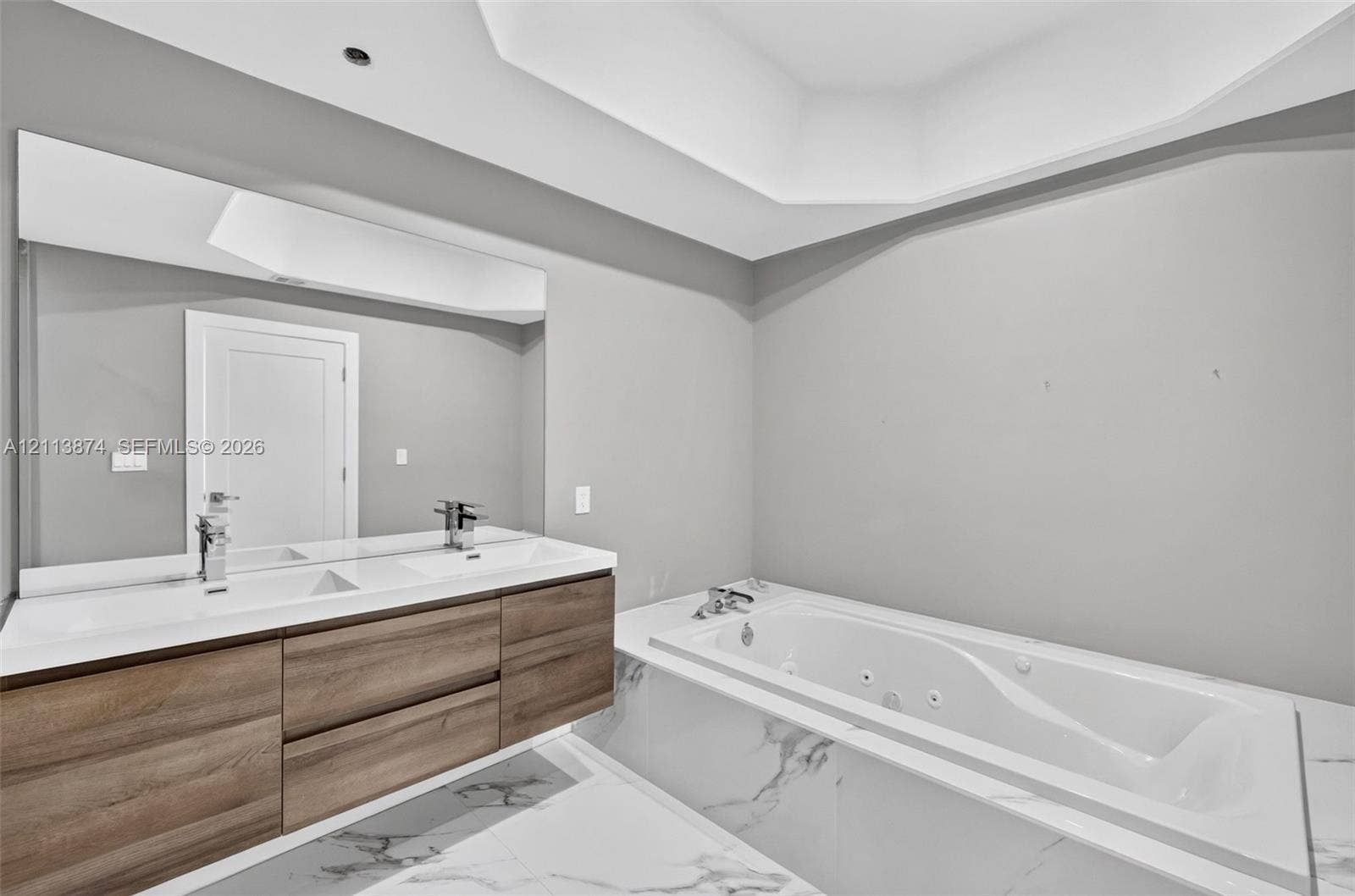 3900 Island Boulevard, #B408, Aventura, Aventura, FL 33160 Floating dual-sink vanity with wood-finish cabinetry, recessed tray ceiling, large wall-spanning mirror, and jetted soaking tub with stone-patterned tile surround