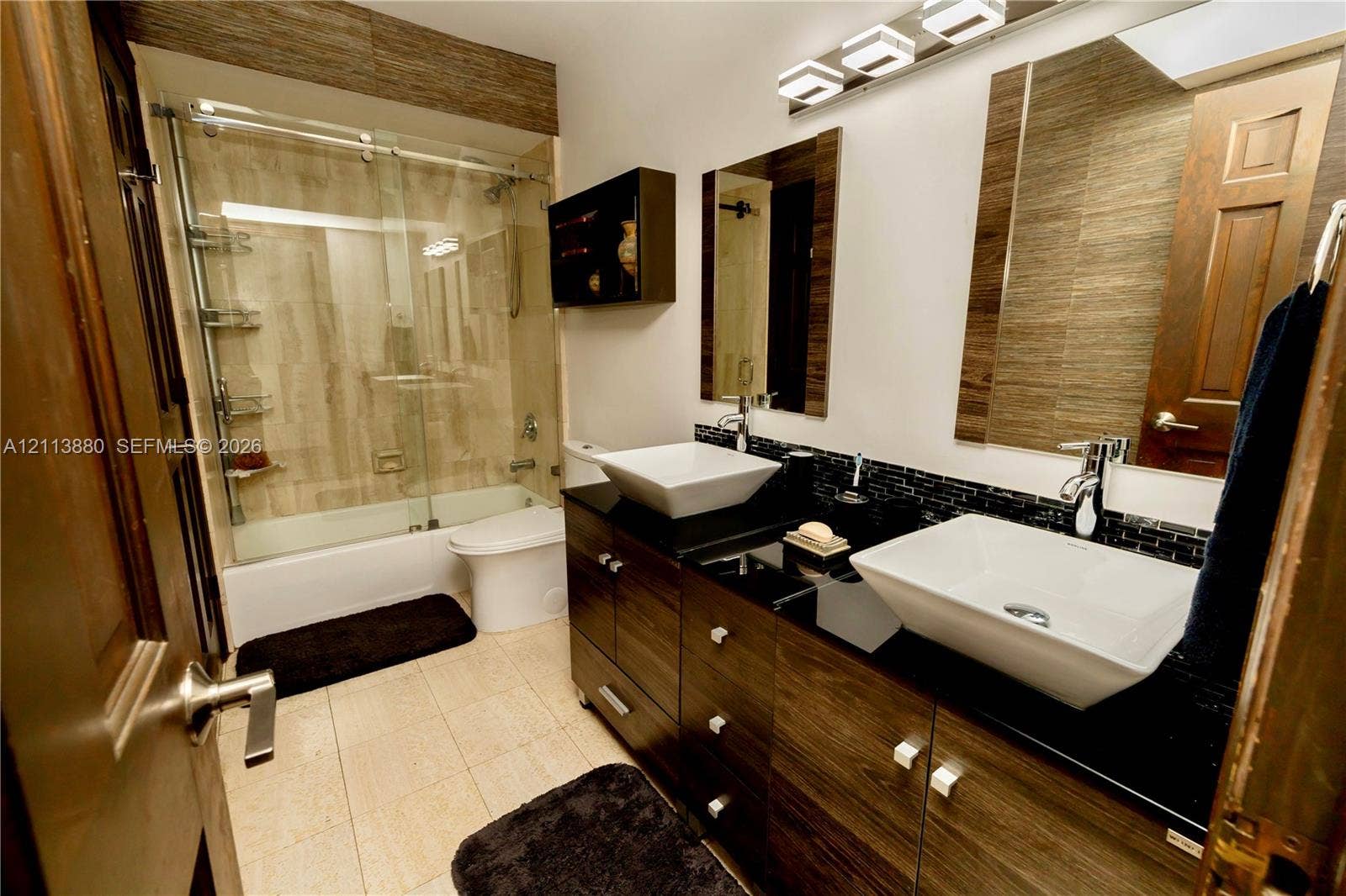 900 Coco Plum Way, Plantation, FL 33324 Dual vessel sinks with polished chrome faucets, black countertop, wood-finish cabinetry, tile-surround shower with sliding glass door, and integrated vanity lighting