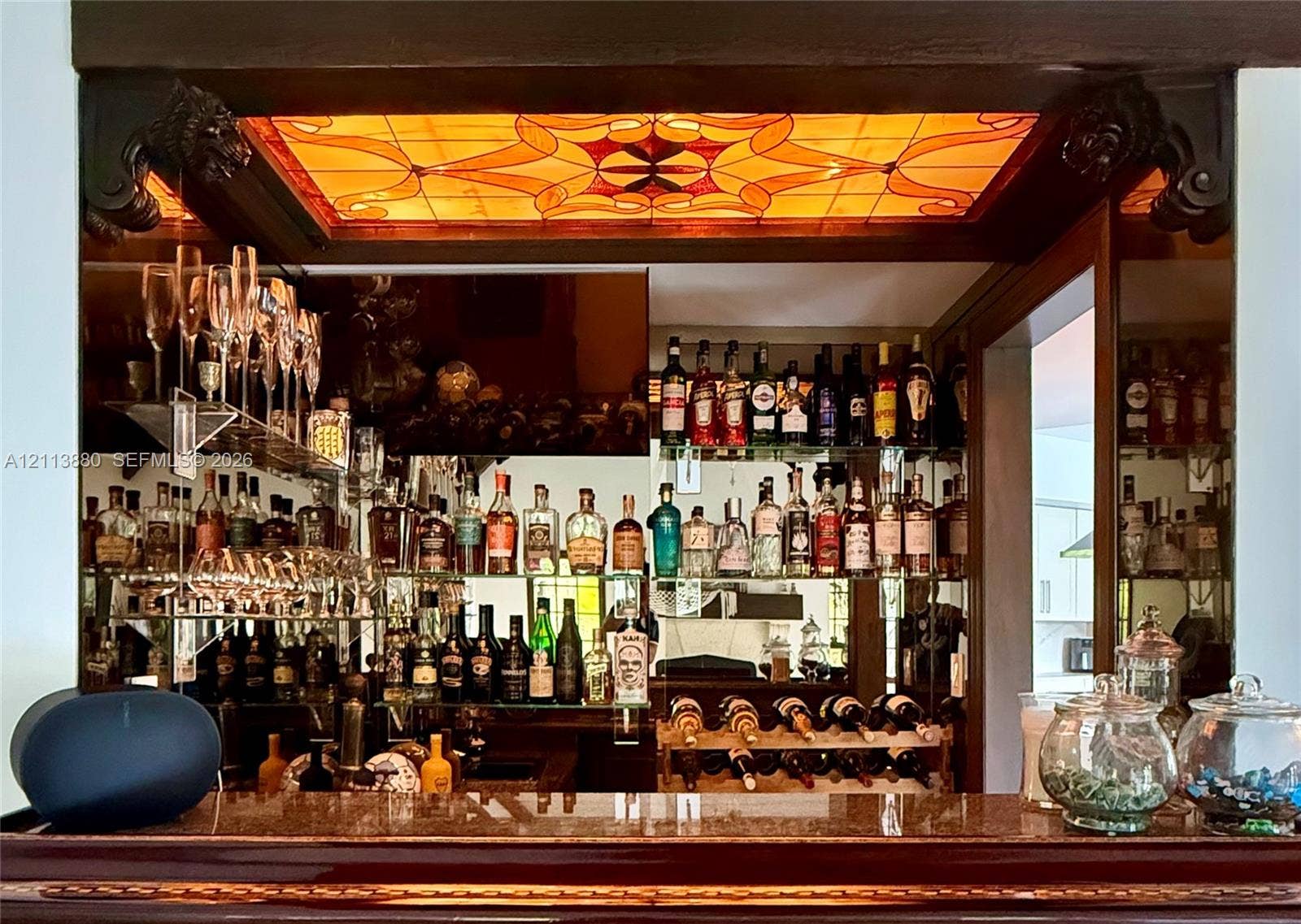 900 Coco Plum Way, Plantation, FL 33324 Built-in bar area featuring a stone countertop, mirrored wall backing, multiple glass shelving tiers, and a decorative amber-hued stained glass ceiling inlay framed by carved wood corbels