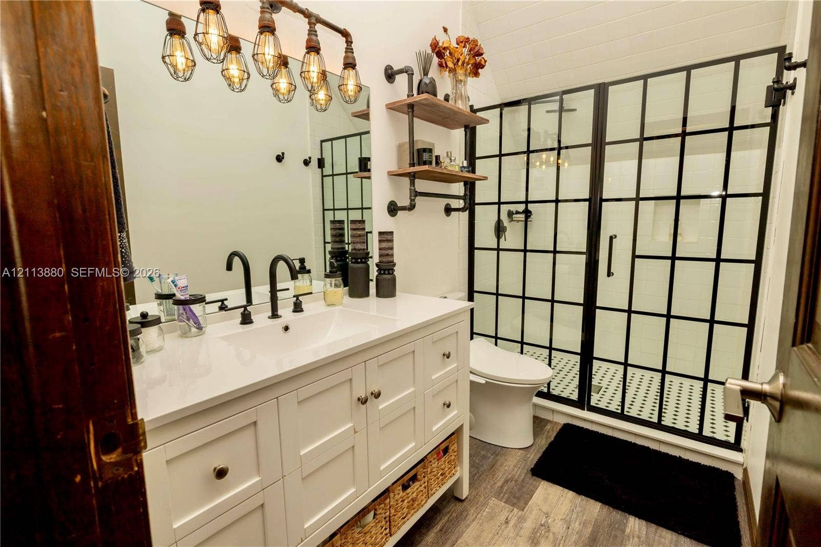 900 Coco Plum Way, Plantation, FL 33324 Bathroom vanity featuring white cabinetry, a solid surface countertop, and matte black widespread faucet, complemented by a walk-in shower with a black grid-frame glass enclosure, industrial-style multi-bulb vanity lighting, and wall-mounted pipe-style shelving
