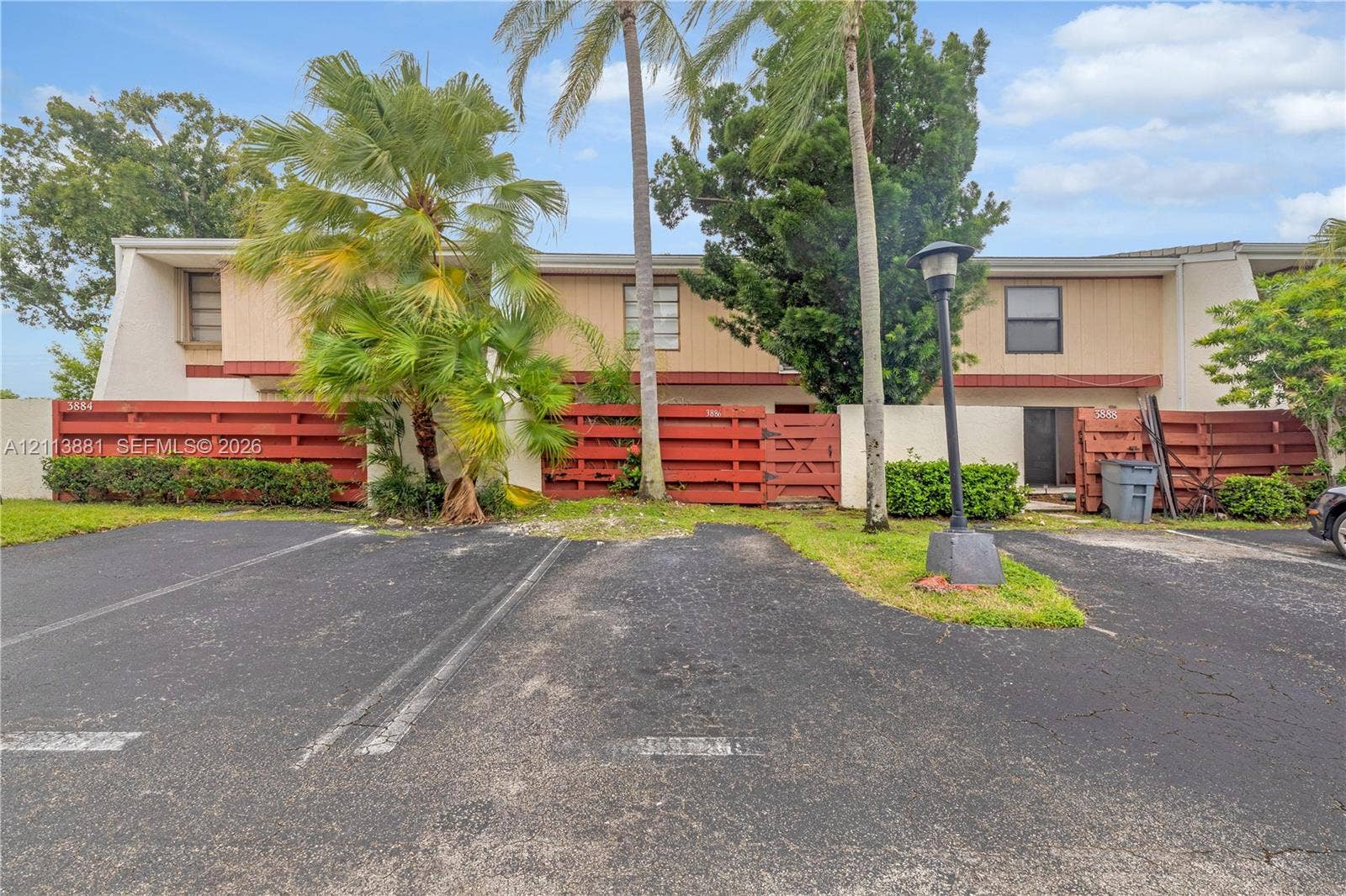 3886 Meadow Lane, #250, Hollywood, FL 33021 Two-story exterior featuring wood-paneled siding, decorative red wood fencing, asphalt parking area, and mature palm landscaping