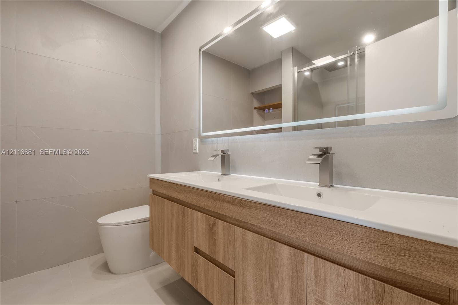 3886 Meadow Lane, #250, Hollywood, FL 33021 Floating wood-finish vanity with dual integrated sinks, wall-mounted metallic faucets, large backlit mirror, and floor-to-ceiling tiled walls