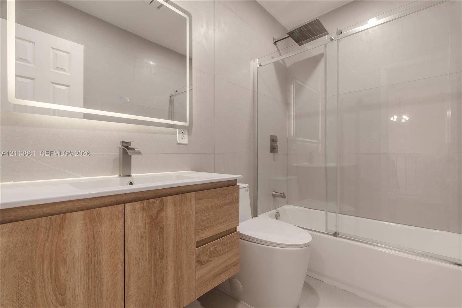 3886 Meadow Lane, #250, Hollywood, FL 33021 Bathroom featuring a wood-finish vanity with a rectangular sink, wall-mounted rectangular LED-lit mirror, and a tub with a glass sliding door enclosure and rain-style shower head